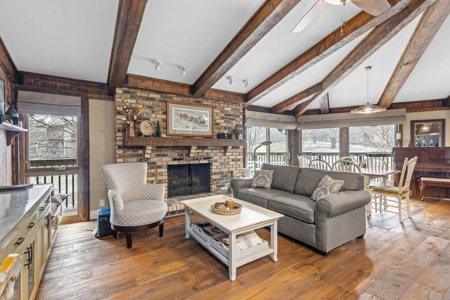 Living room with brick fireplace, gray sofa, white armchair, wooden coffee table, large windows with snow outside, and a dining area with a round table and chairs.