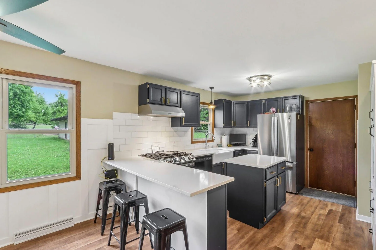 Kitchen with black cabinets, white countertops, stainless steel appliances, window with outdoor view, and a small breakfast bar area with three black bar stools.
