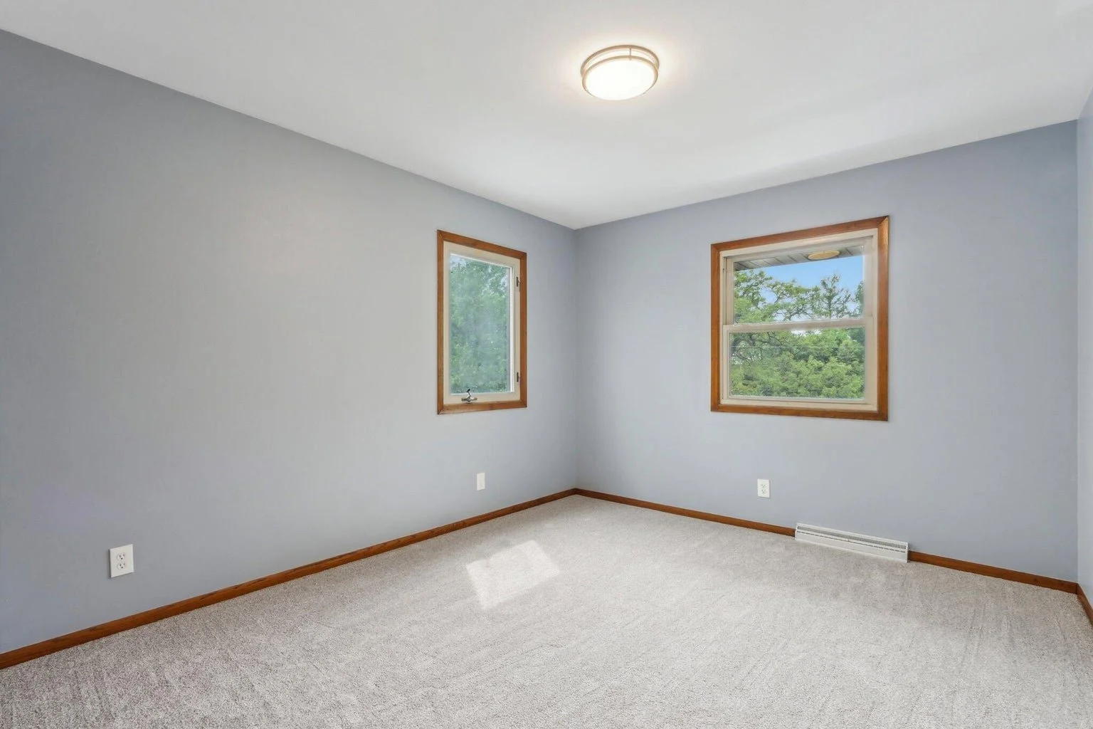Empty room with two windows with wooden frames, light blue walls, beige carpet, and a ceiling light fixture.