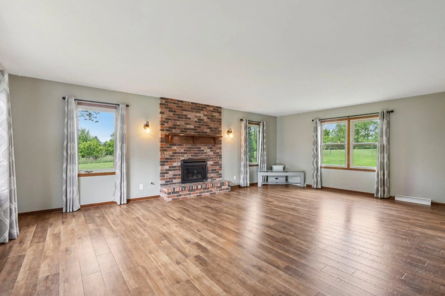 Empty living room with hardwood floors, brick fireplace, large windows with curtains, and a small white shelf