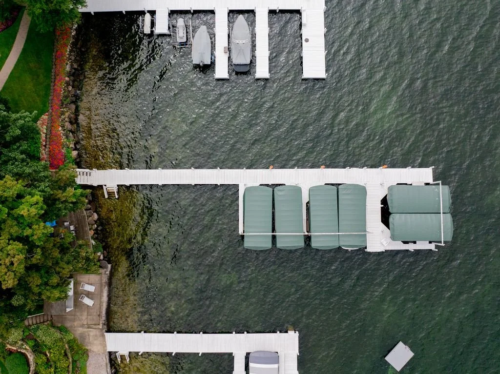 An aerial view of a lakeside dock with boat slips, boats, a narrow walkway, and surrounding greenery.