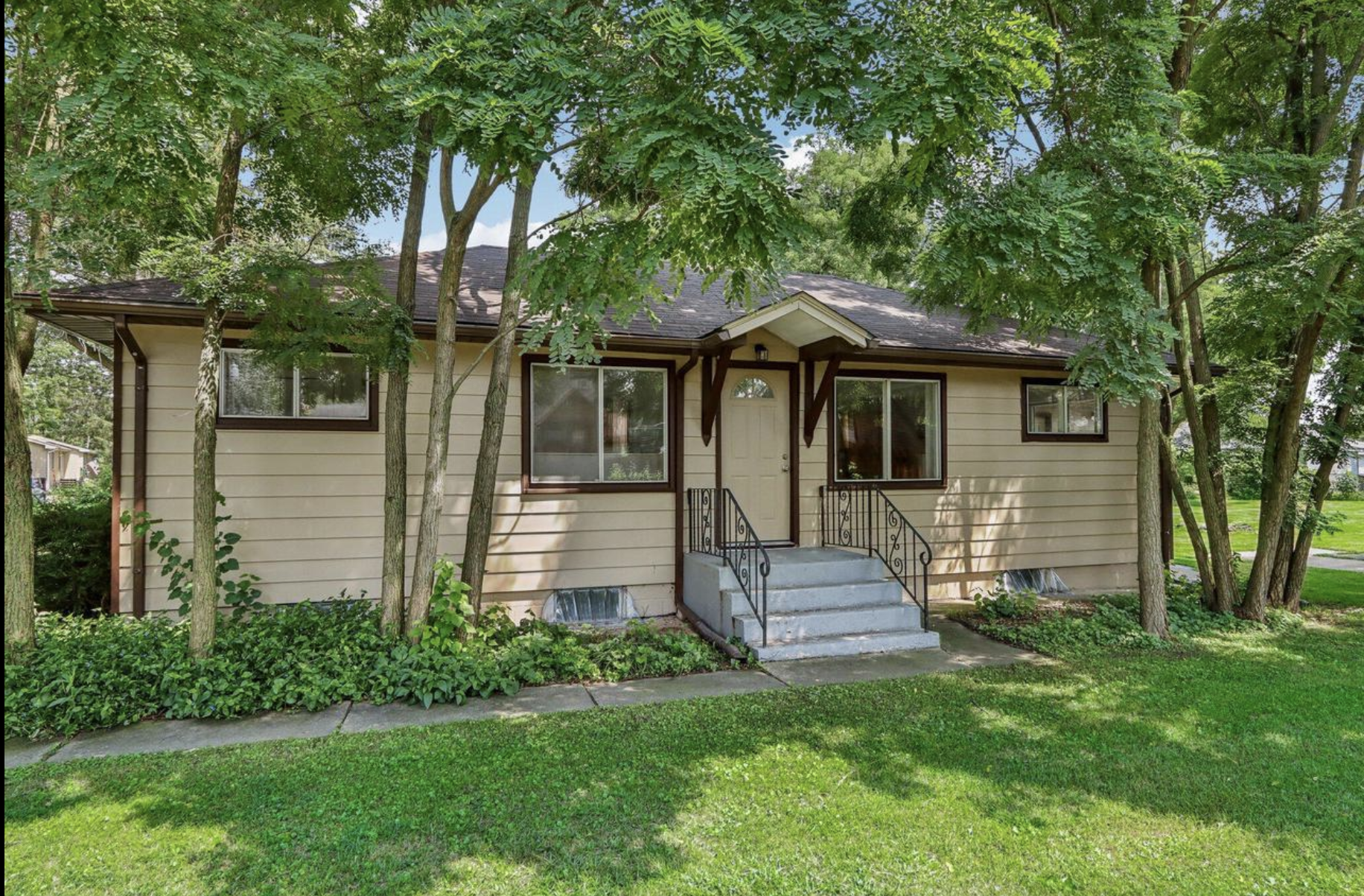 A beige house with a gabled roof, surrounded by tall trees, with a small porch and steps leading to the front door, and landscaped with bushes and grass.