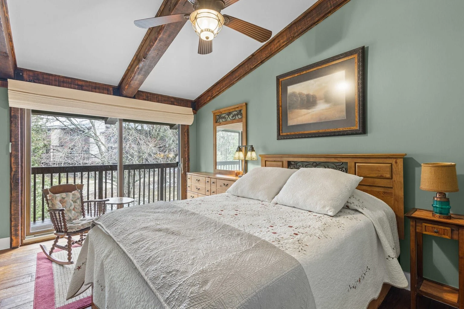 Cozy bedroom with wood-beamed ceiling, large sliding glass door leading to a balcony, and a bed with floral quilt and white pillows.