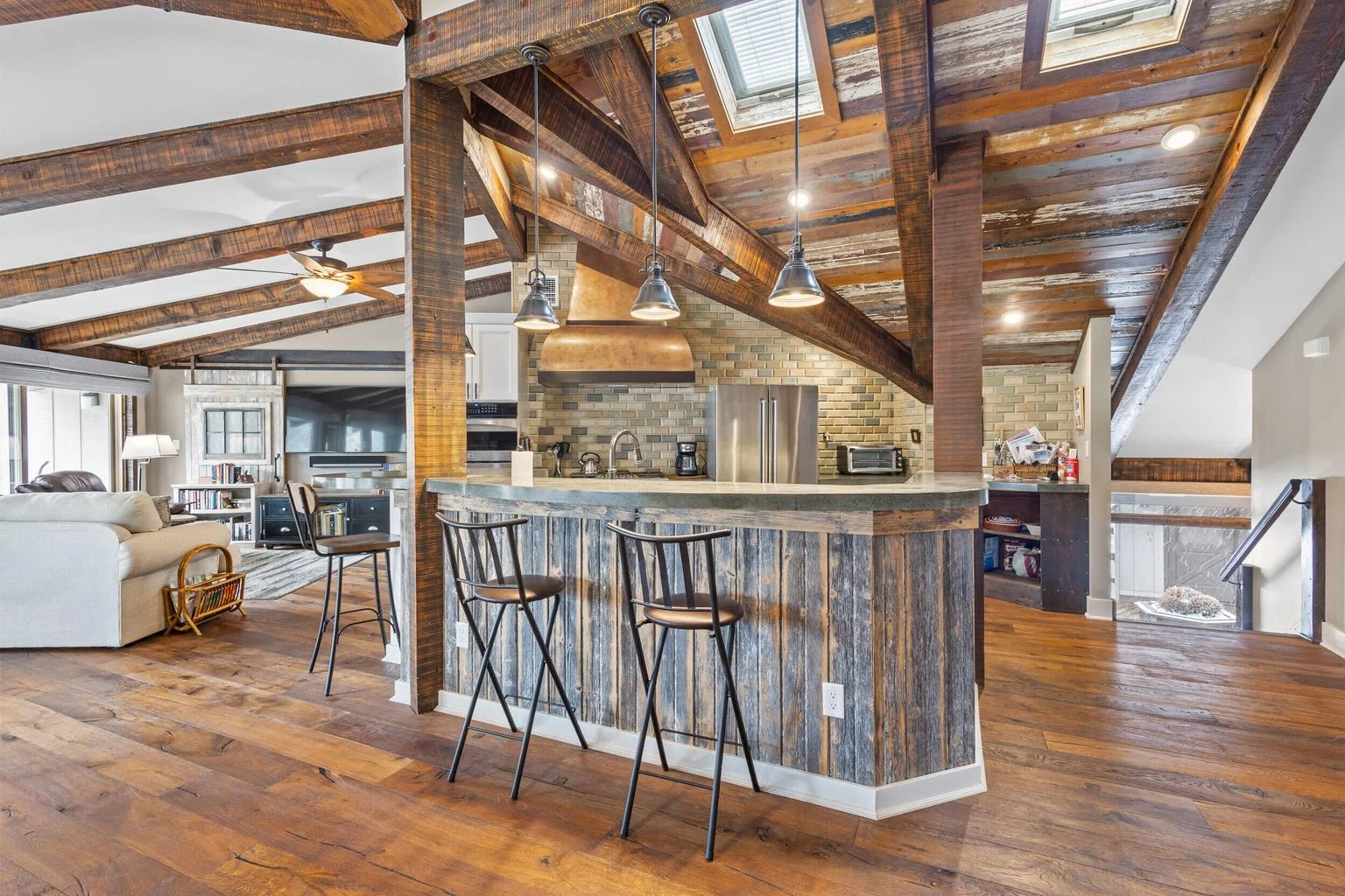 Open-concept living area with a rustic wooden kitchen bar, metal bar stools, and a cozy living room with a beige sofa, a bookcase, and a TV, featuring a wood-paneled ceiling, hardwood floors, and a staircase.