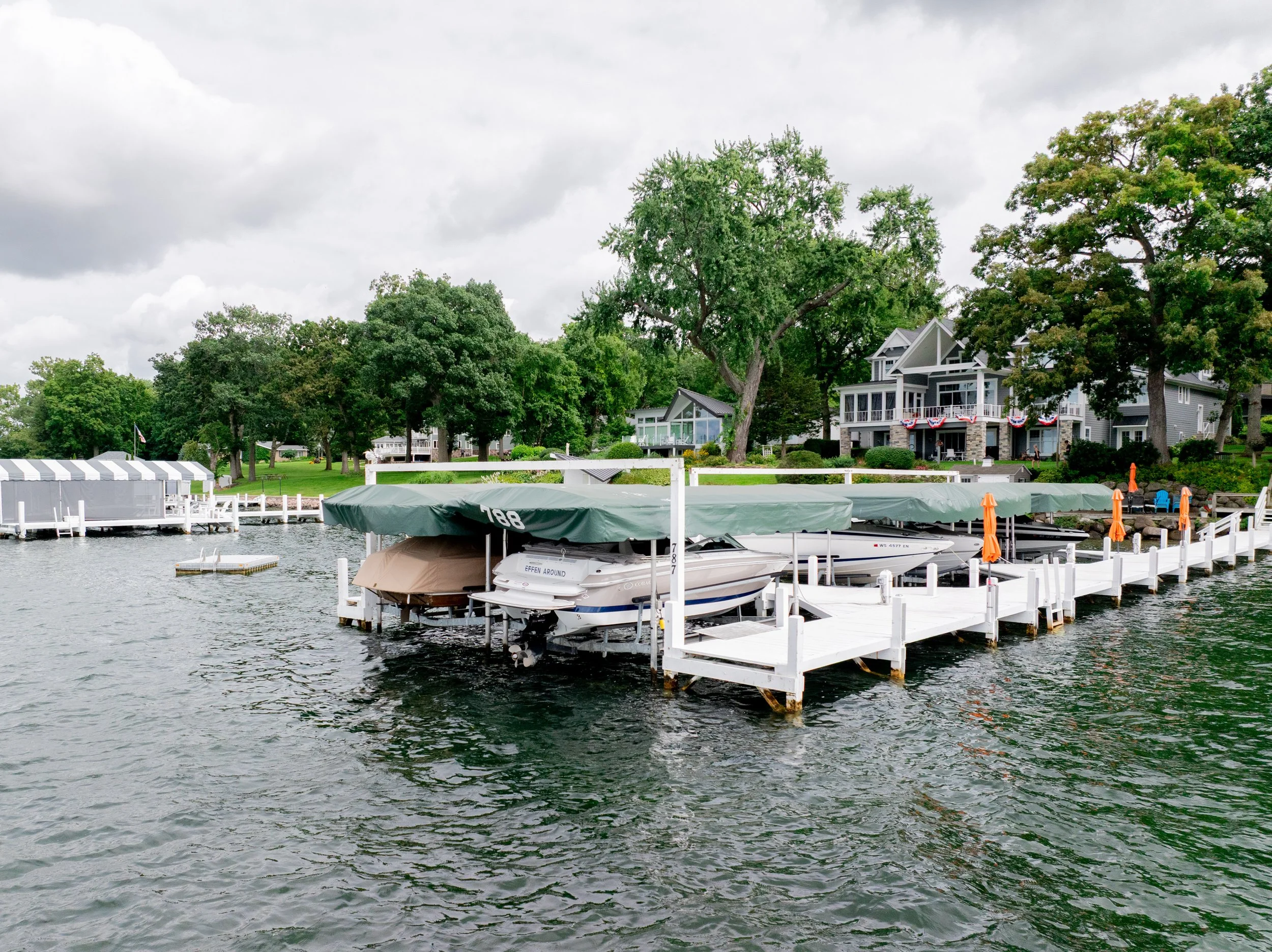 A lakeside view with boat docks and boats under a cloudy sky, surrounded by green trees and residential houses.