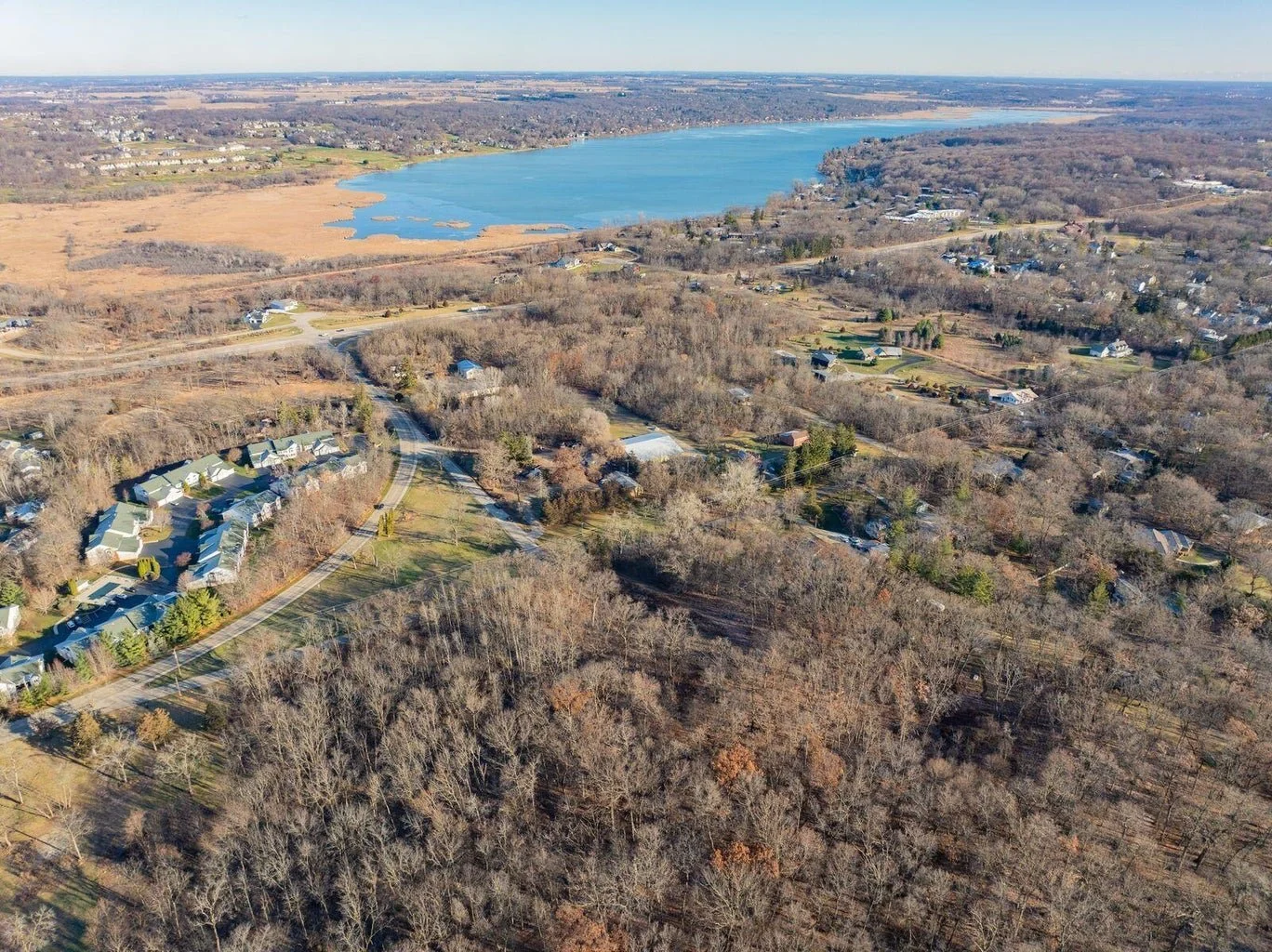 Aerial view of a lakeside residential area with houses, trees, and a large lake in the background, with some open fields nearby.