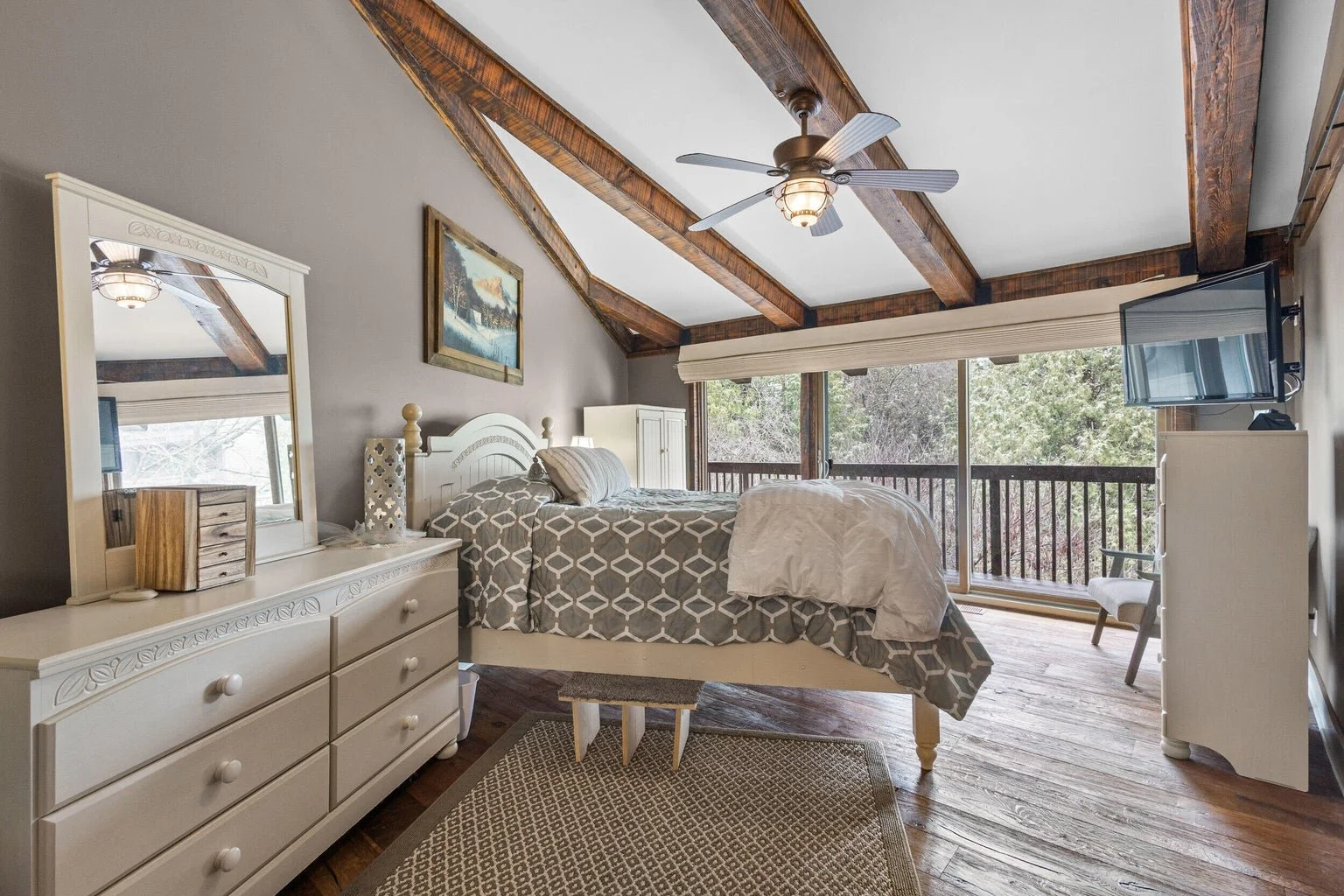 A cozy bedroom with a sloped ceiling and exposed wooden beams, featuring a white dresser with a mirror, a bed with patterned gray and white bedding, a small gray ottoman at the foot of the bed, a window with a view of trees, and a flat-screen TV on a