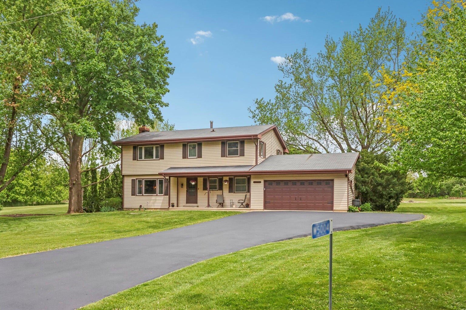 Two-story house with beige siding, brown roof, and attached garage situated on a lush green lawn surrounded by trees, with a driveway leading to the garage and a street sign in the foreground.
