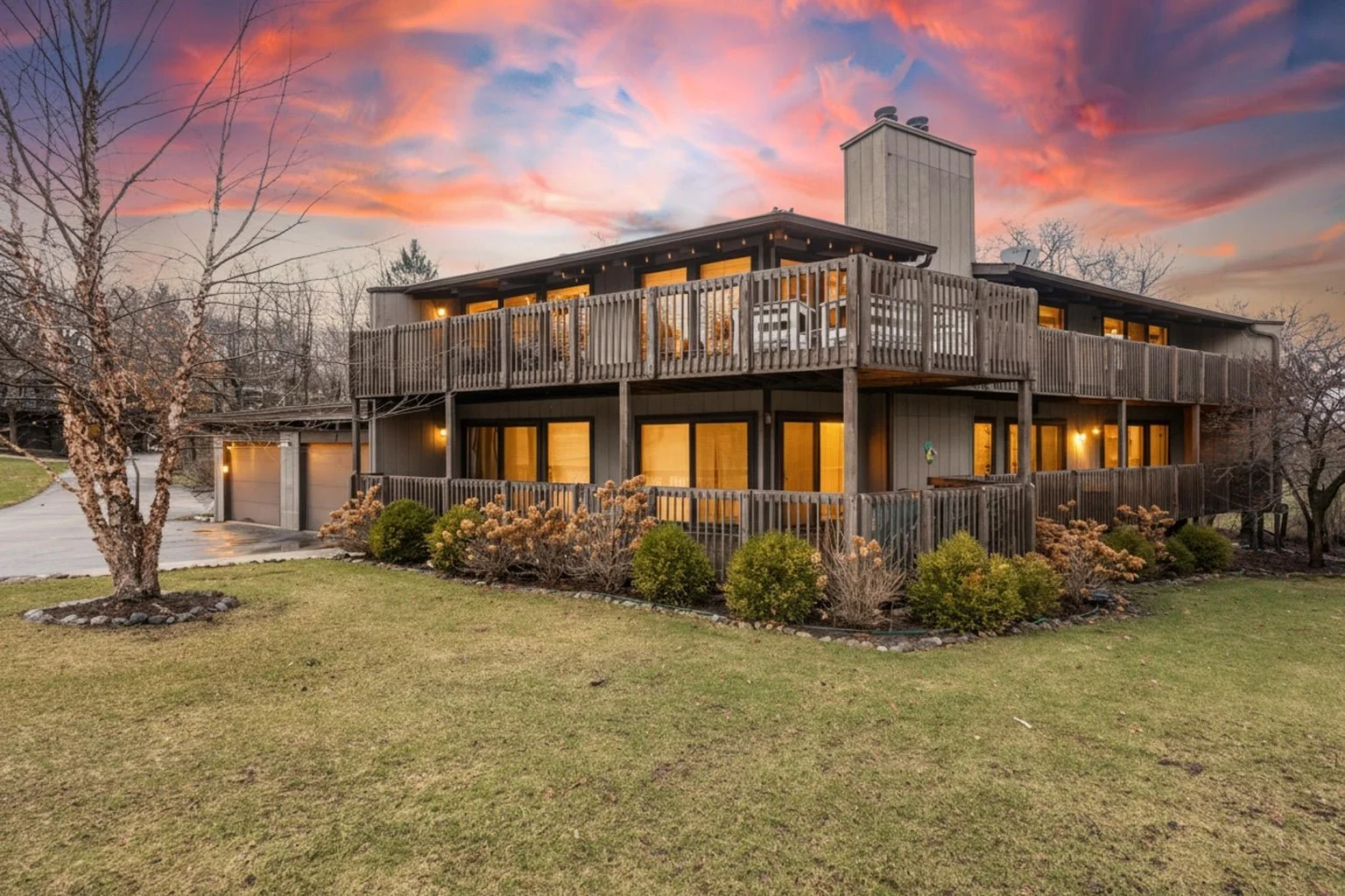 A two-story wooden house with large windows, a balcony on the upper level, surrounded by a landscaped lawn with shrubs and trees, at sunset with a colorful sky.