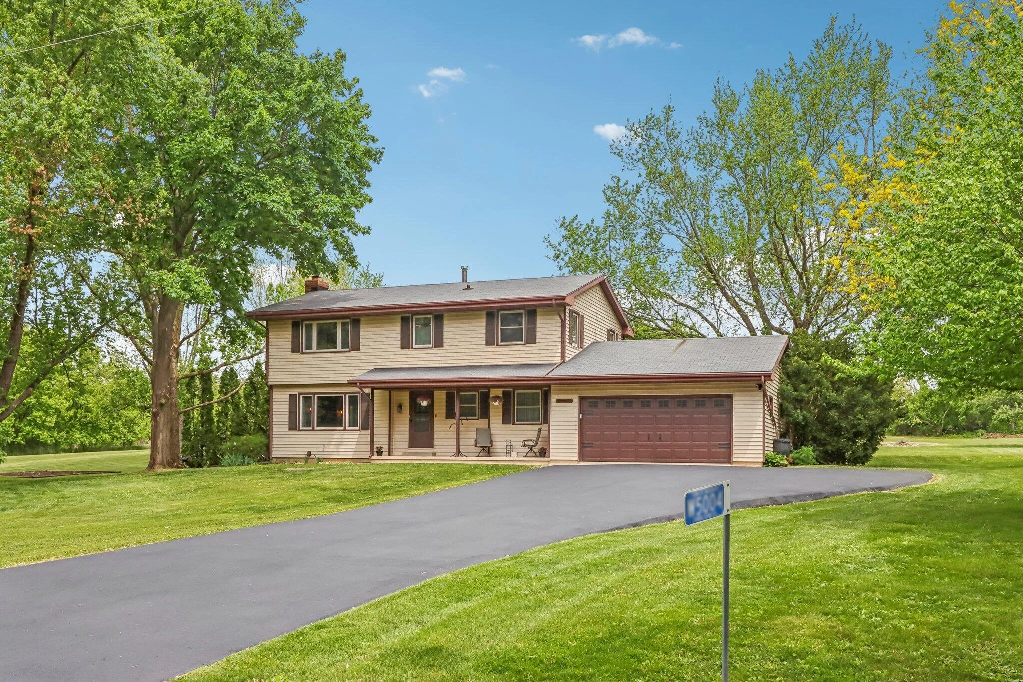 A two-story house with beige siding, brown window shutters, and a brown garage door, surrounded by green grass and large trees, with a clear blue sky overhead.