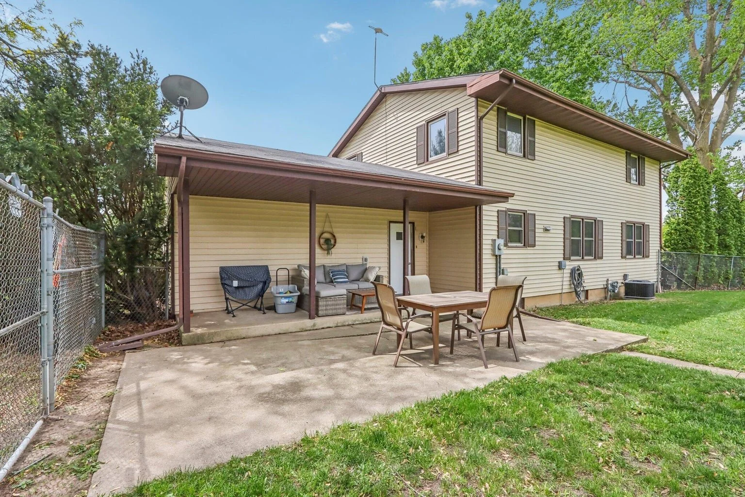 Backyard patio area with outdoor furniture, including a table and chairs, on a concrete slab next to a beige house with brown trim and window shutters. Green lawn and trees in the background.