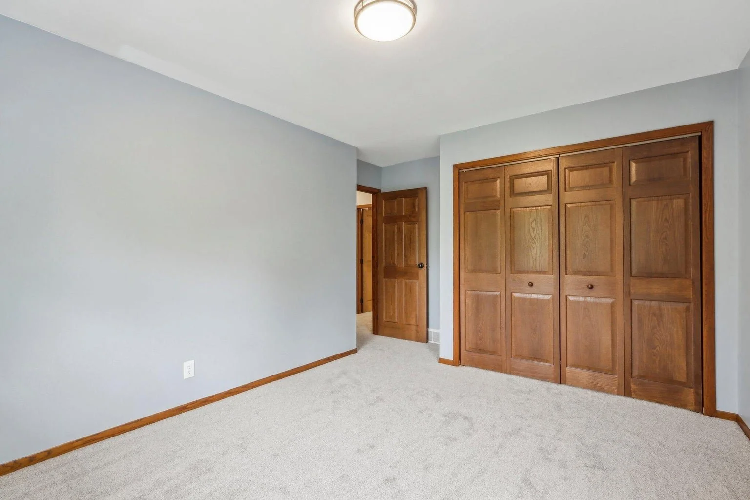 Empty bedroom with light gray walls, beige carpet, a wooden closet with folding doors, and an open wooden door leading to another room.
