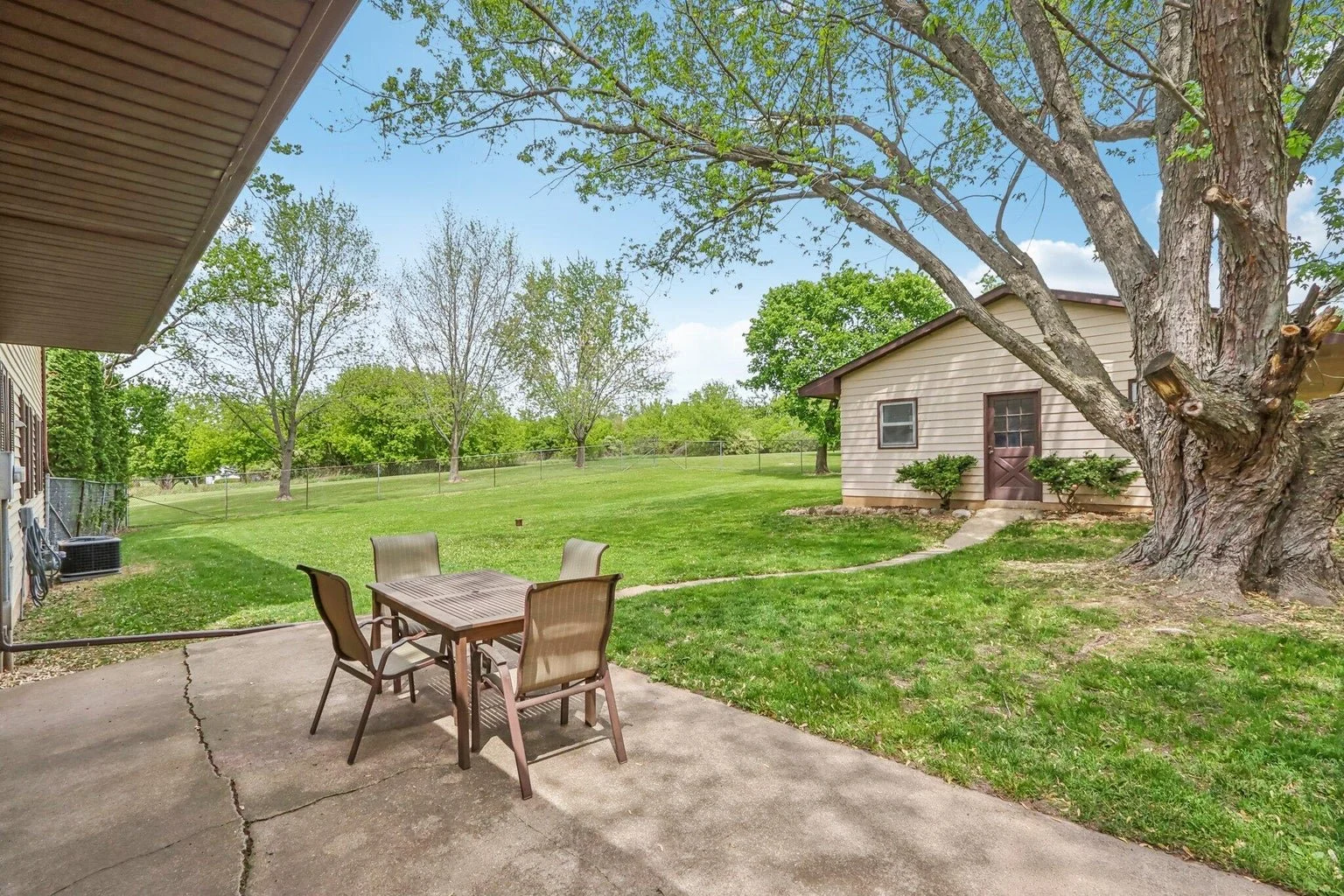 Backyard patio with a table and chairs, large tree, green lawn, small shed, and trees in the background under a partly cloudy sky.