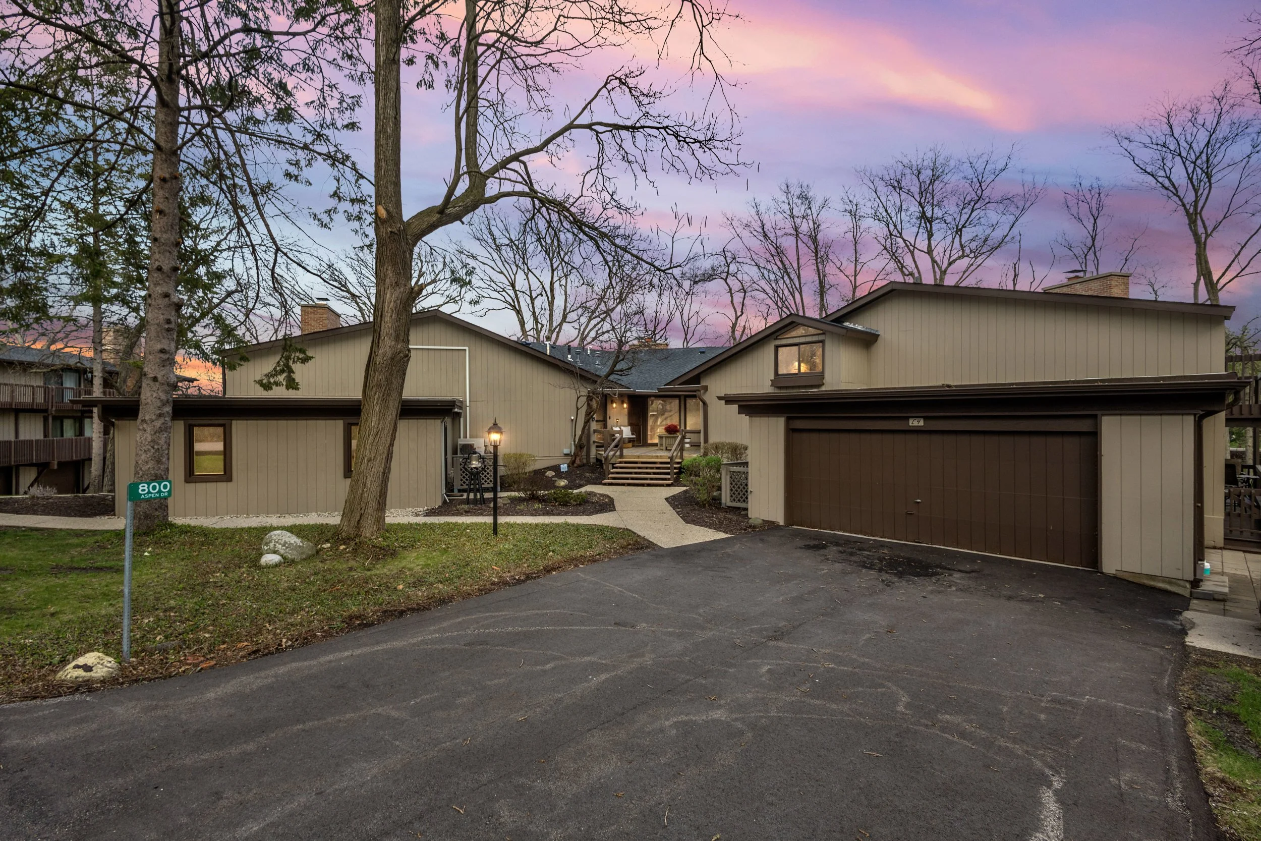 A modern two-story house with beige siding, large front windows, and a brown garage door. The house has a small front porch with steps, outdoor lighting, and a paved driveway. The front yard features a lawn, a small garden, and a street sign that reads '800 Aspen Dr'. Tall trees with bare branches are visible against a colorful sunset sky with pink and purple clouds.