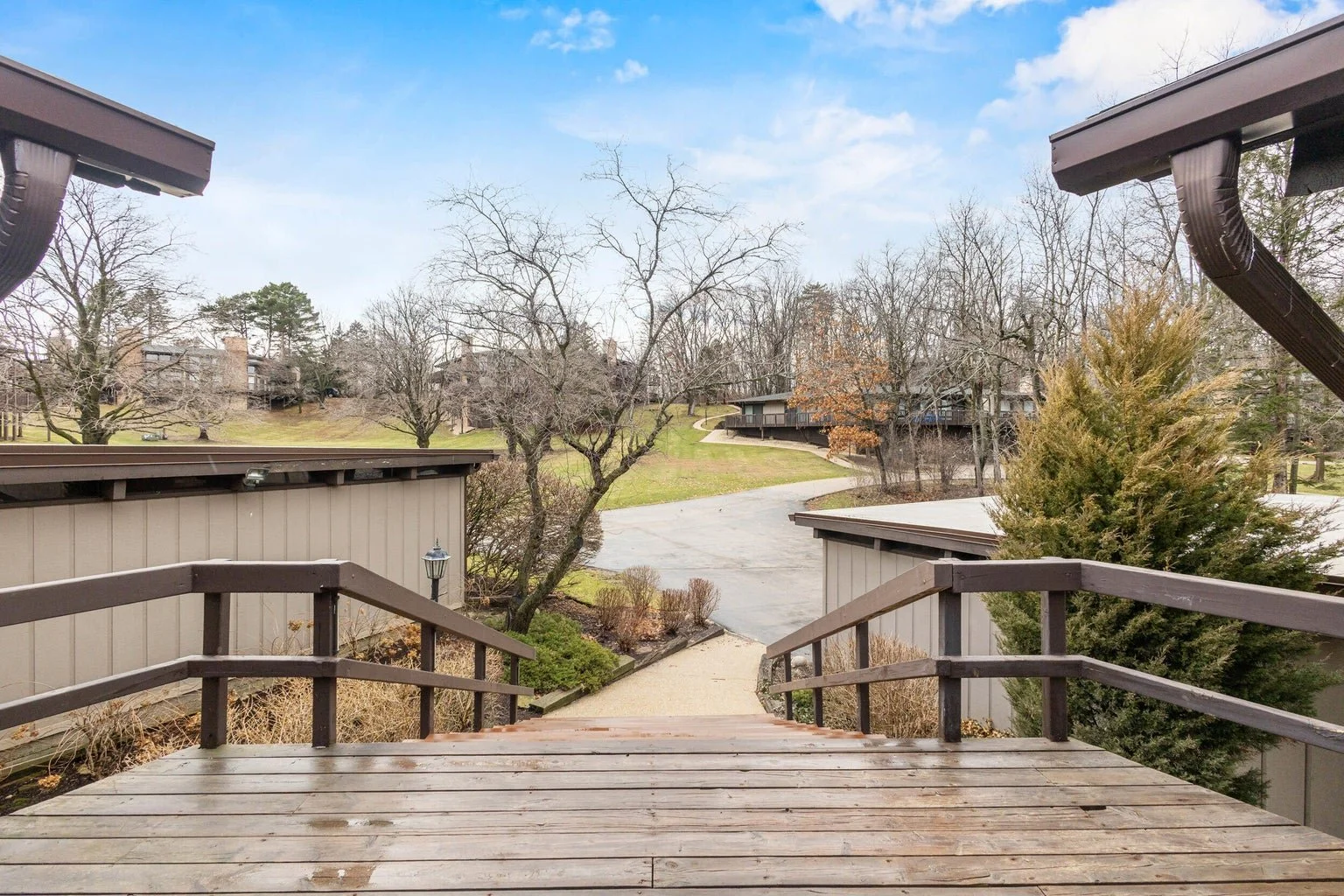 View from a wooden deck looking out over a paved path in a residential area with leafless trees, grassy areas, and houses in the background on a partly cloudy day.