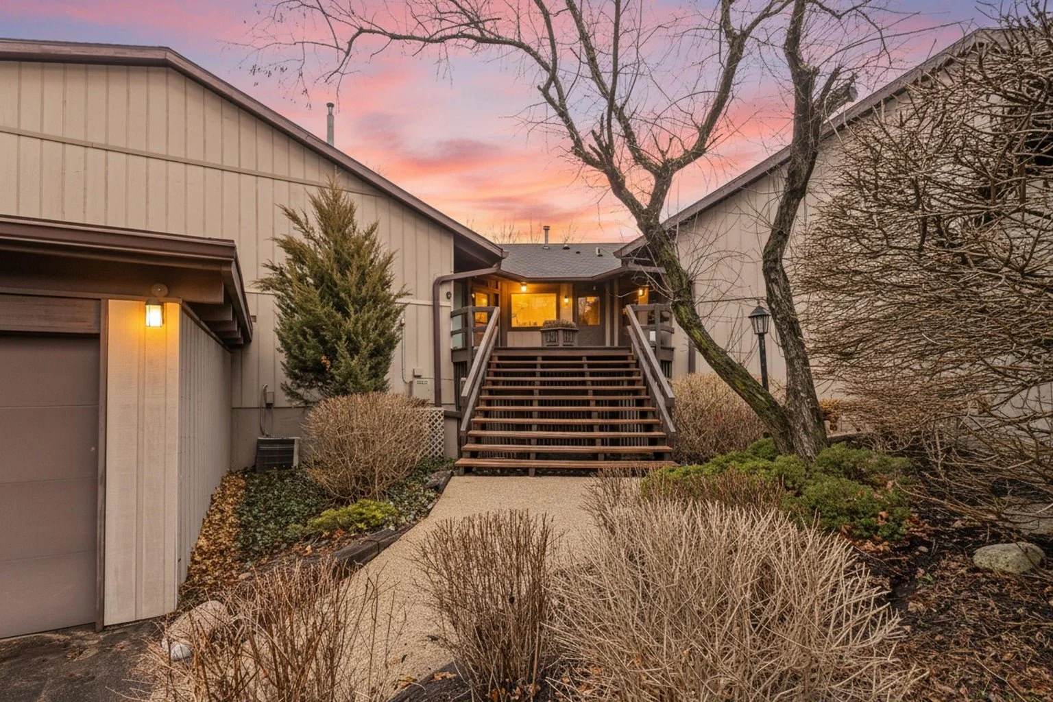 A house with a deck at sunset. The house has beige exterior walls and a sloped roof. There is a staircase leading up to the deck, with its lights on. The yard has various leafless bushes and trees, and the sky is painted with pink and purple clouds.