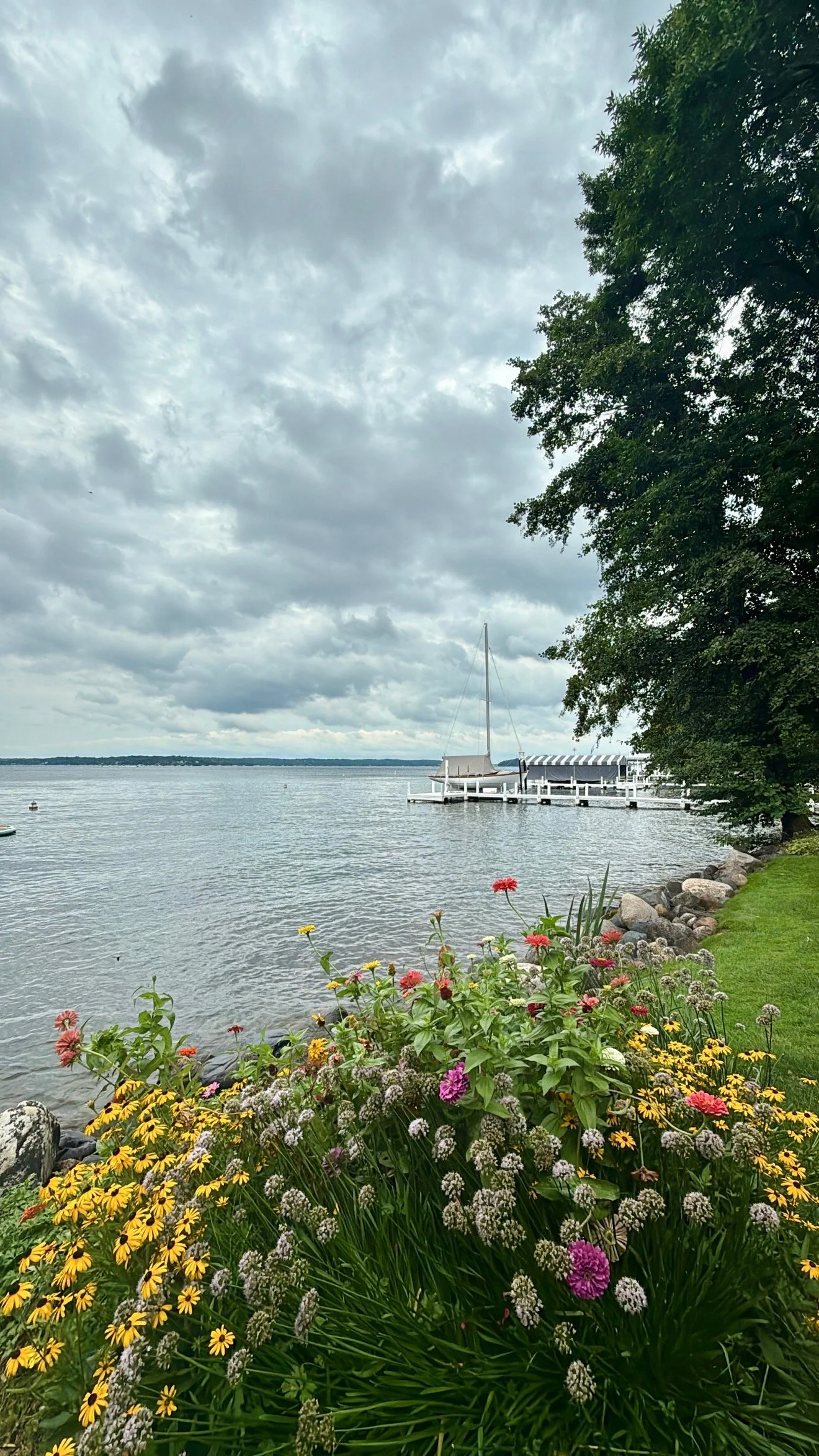 A lakeside scene with colorful flowers in the foreground, a dock with a sailboat, and a cloudy sky overhead.