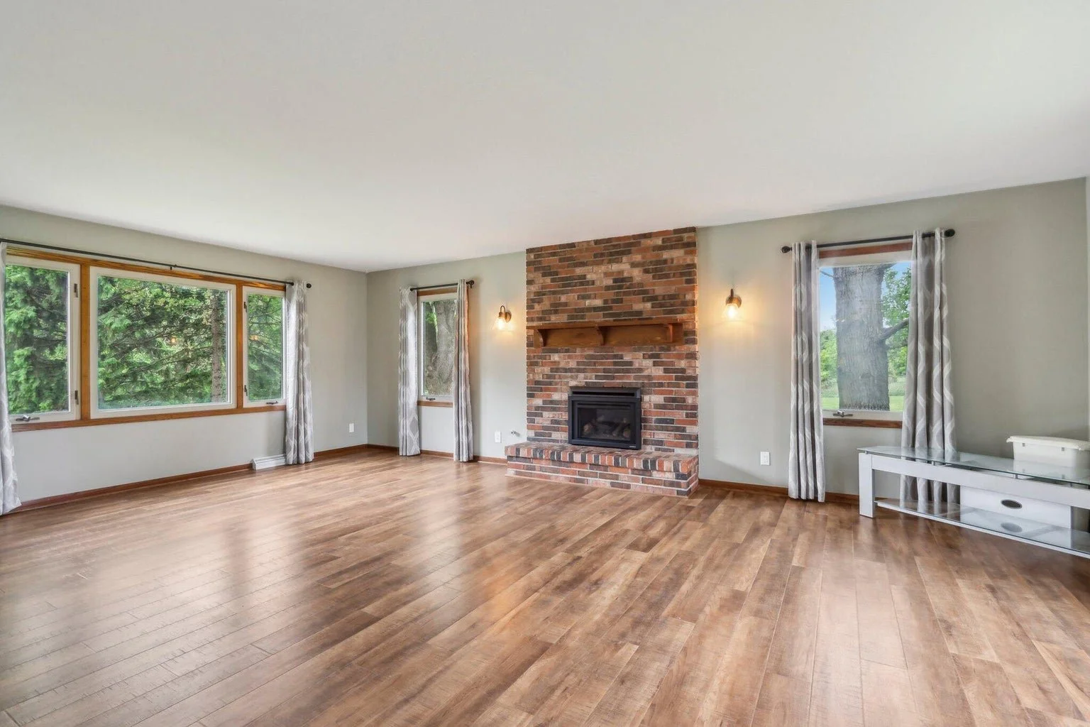 Living room with hardwood floors, brick fireplace, large windows, and curtains.