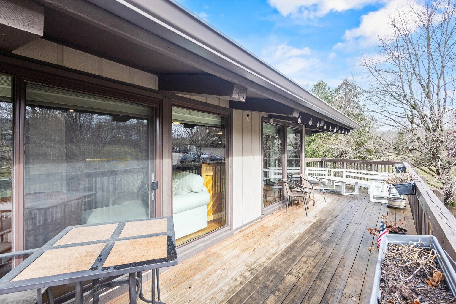 A spacious outdoor balcony with wooden flooring, chairs, a bench, and potted plants, overlooking trees and neighboring houses.