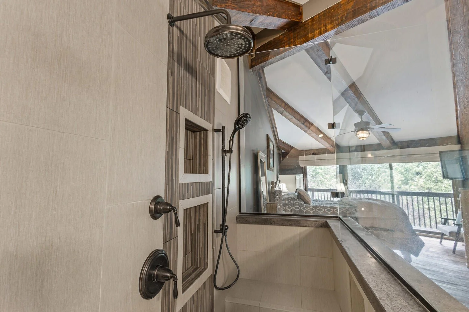 Bathroom shower area with a showerhead, handheld shower wand, built-in niche, and a large mirror overlooking a bedroom with a balcony