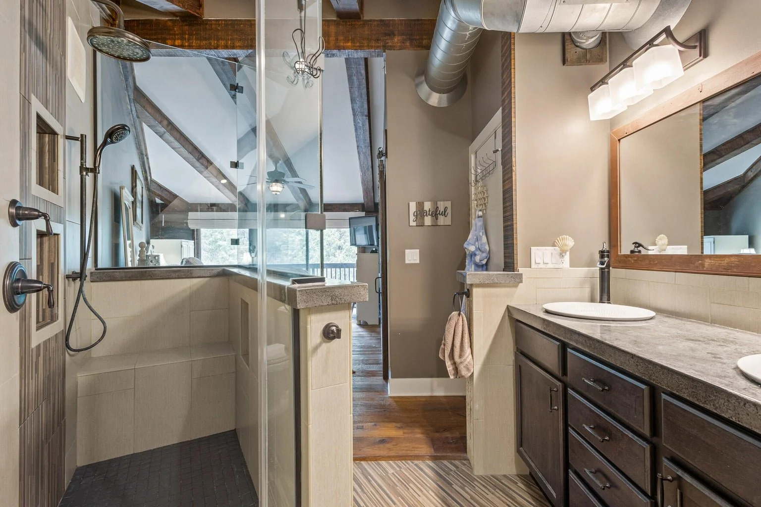 Bathroom with dual sinks, a large mirror, wooden cabinetry, beige tiled walls, a glass shower enclosure, and a window with a view of trees outside.