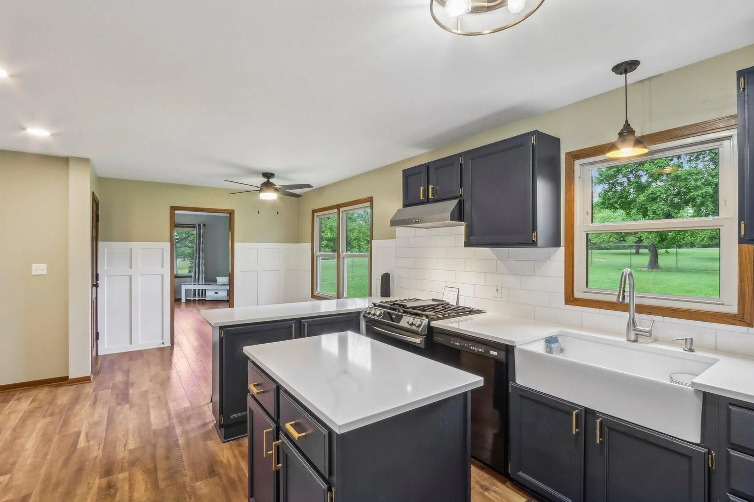 Kitchen with navy blue cabinets, white countertops, white subway tile backsplash, white farmhouse sink, window overlooking green yard, and wooden trim around the window. Open doorway leads to a room with hardwood floor and window.