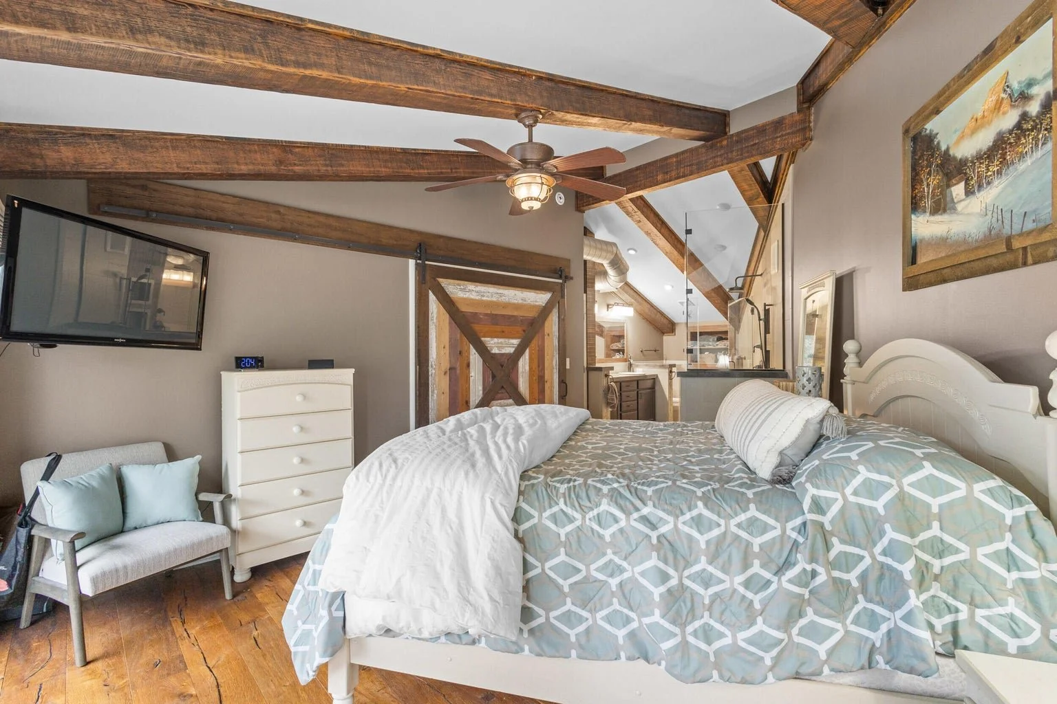 Bedroom with a bed, white dresser, small chair, and a mounted TV, featuring exposed wooden beams and a view into the kitchen area.