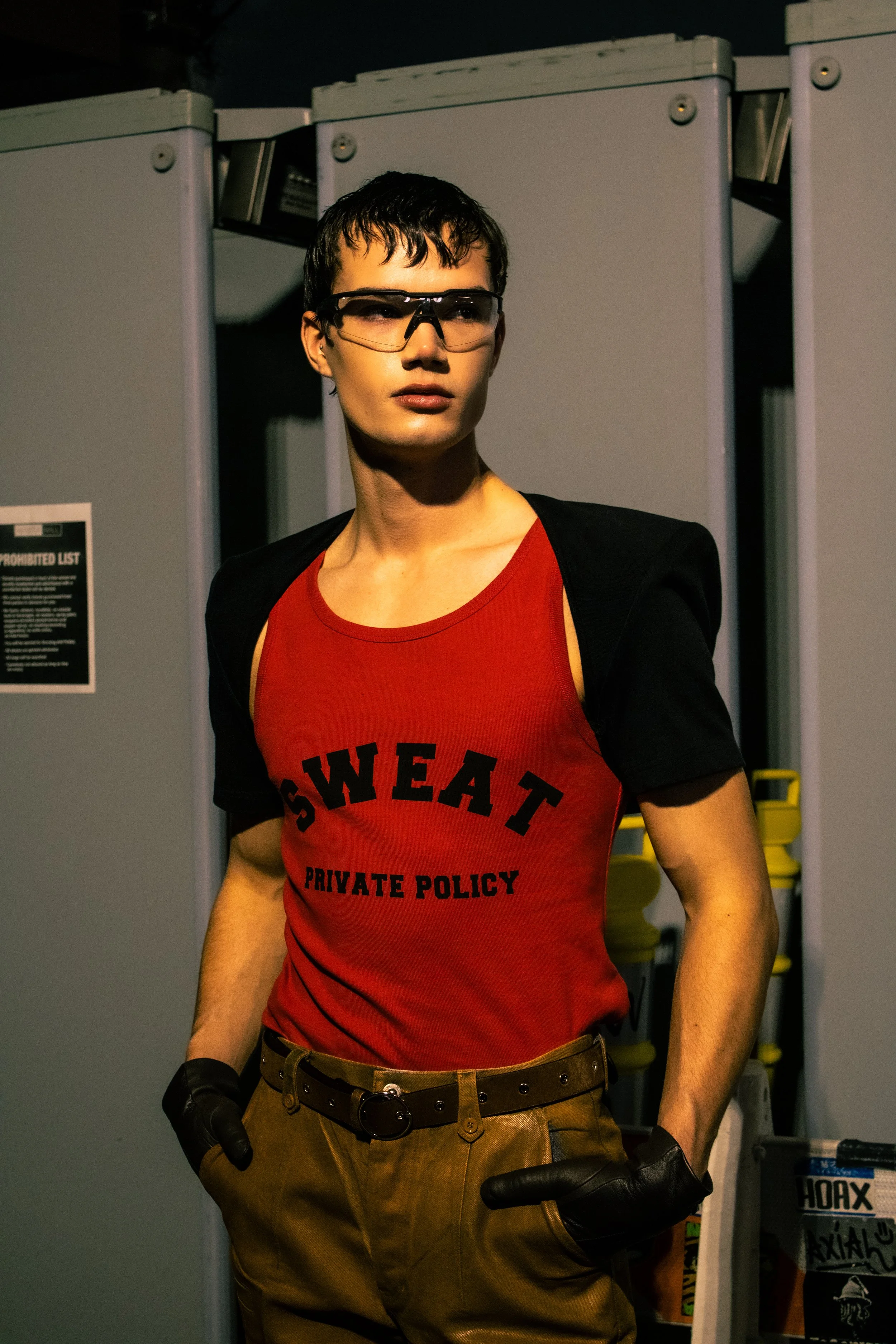 A person standing with hands in pockets, wearing glasses, a red tank top with black text, black gloves, and brown pants, in front of gray lockers.