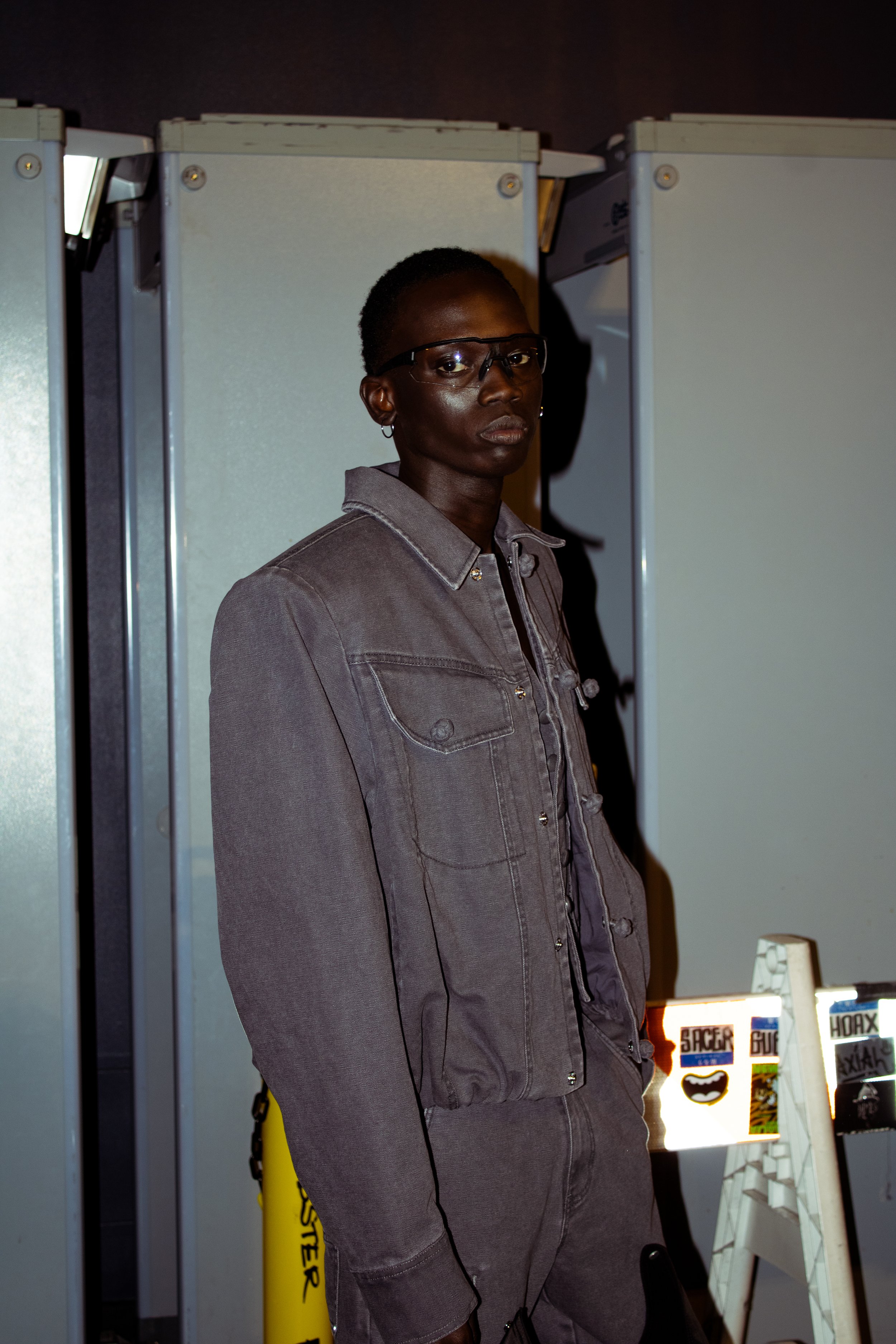 A young man with short hair, wearing glasses and a gray denim jacket, standing indoors in front of a grey background.