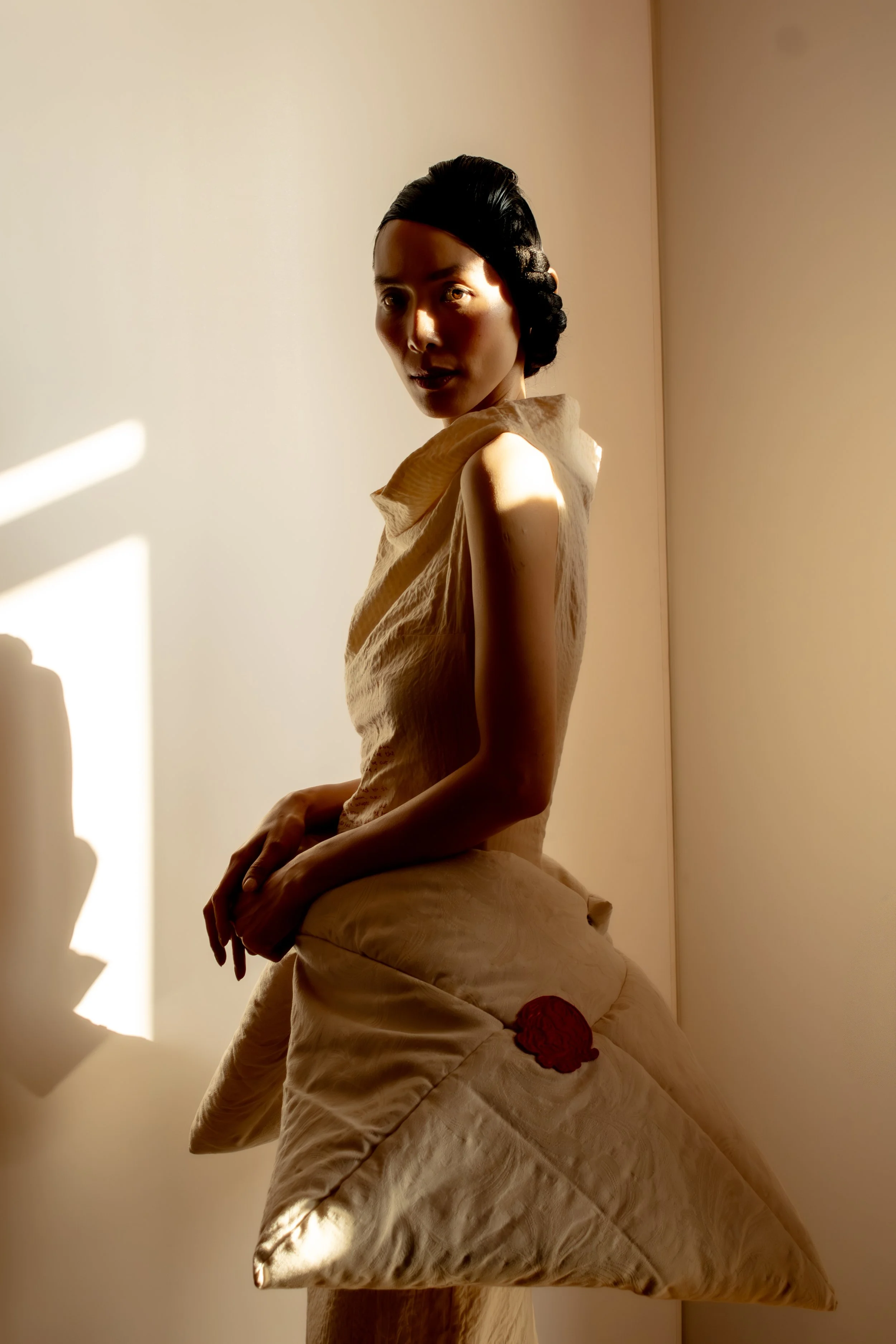 A woman wearing a bold dress with embroidered detail, in front of a plain wall with sunlight casting shadows.