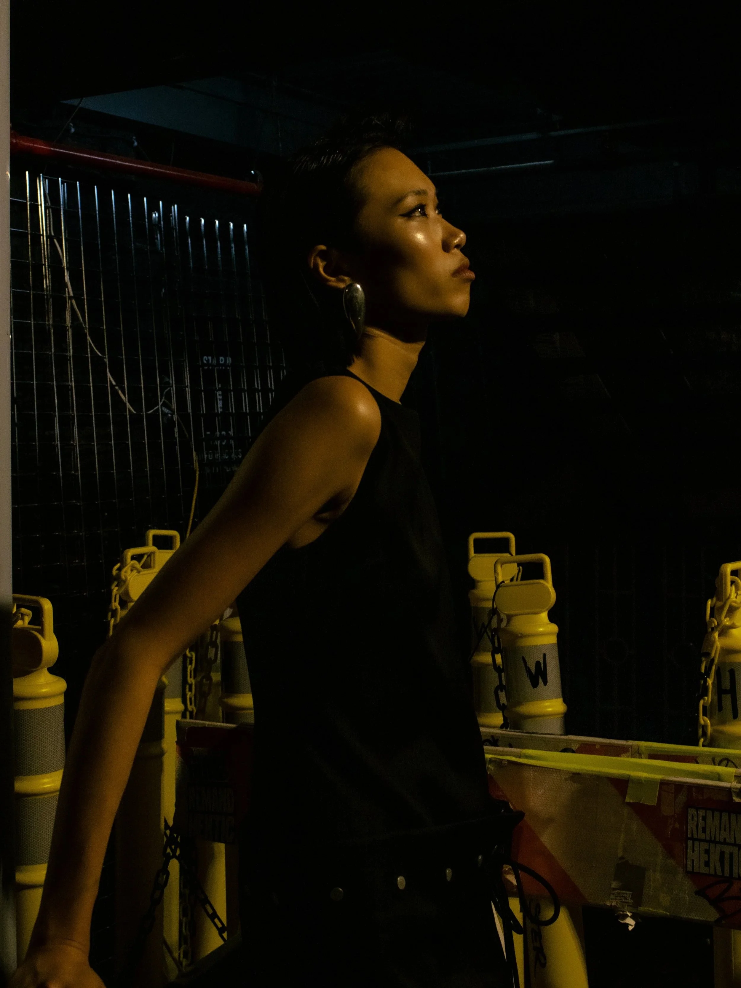 A woman with short dark hair wearing a sleeveless black top and large earrings, standing in a dark space with yellow safety barriers and chains.