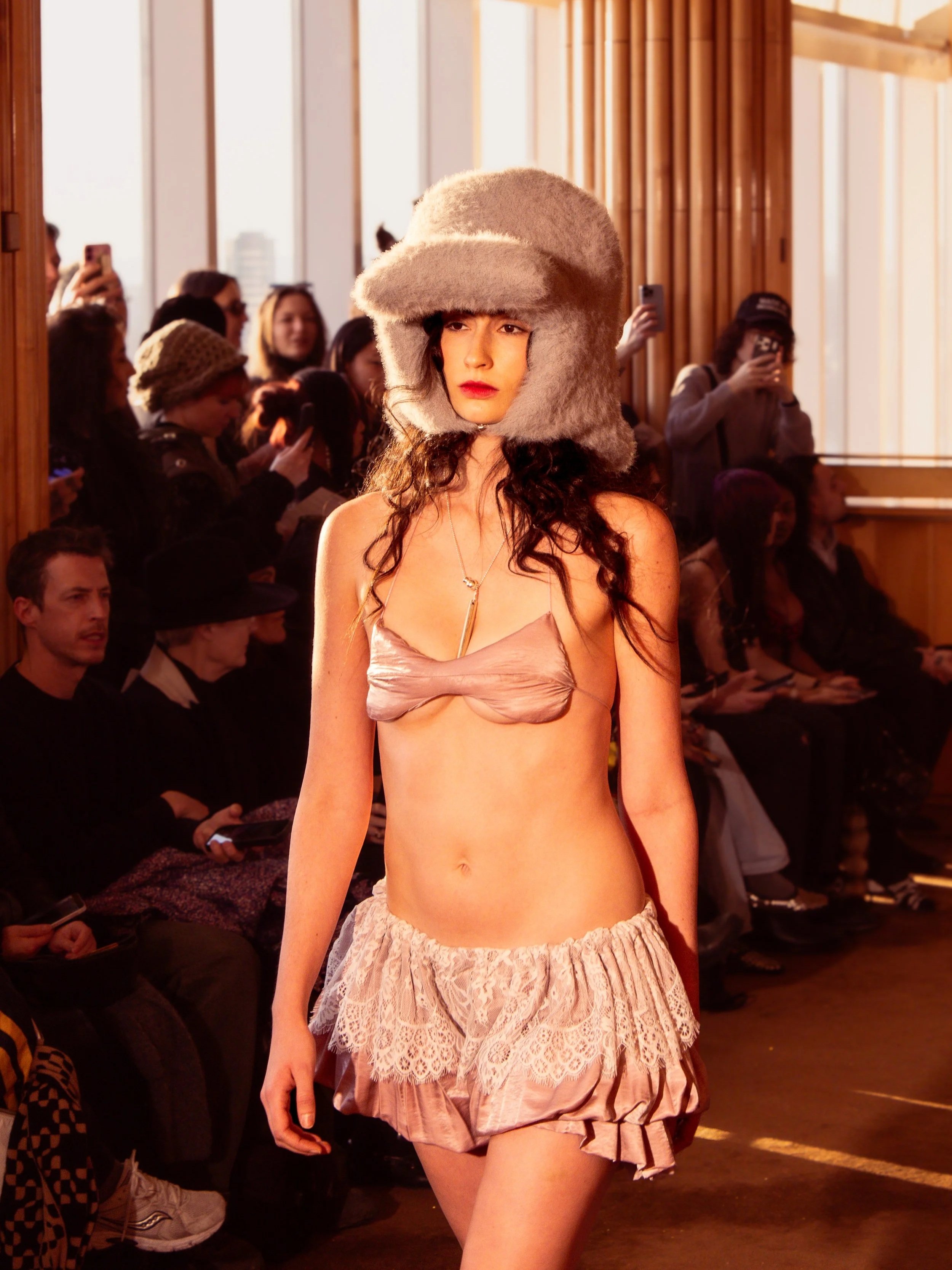 Model walking on runway wearing a large fuzzy hat and minimal clothing, audience watching and taking photos in background.