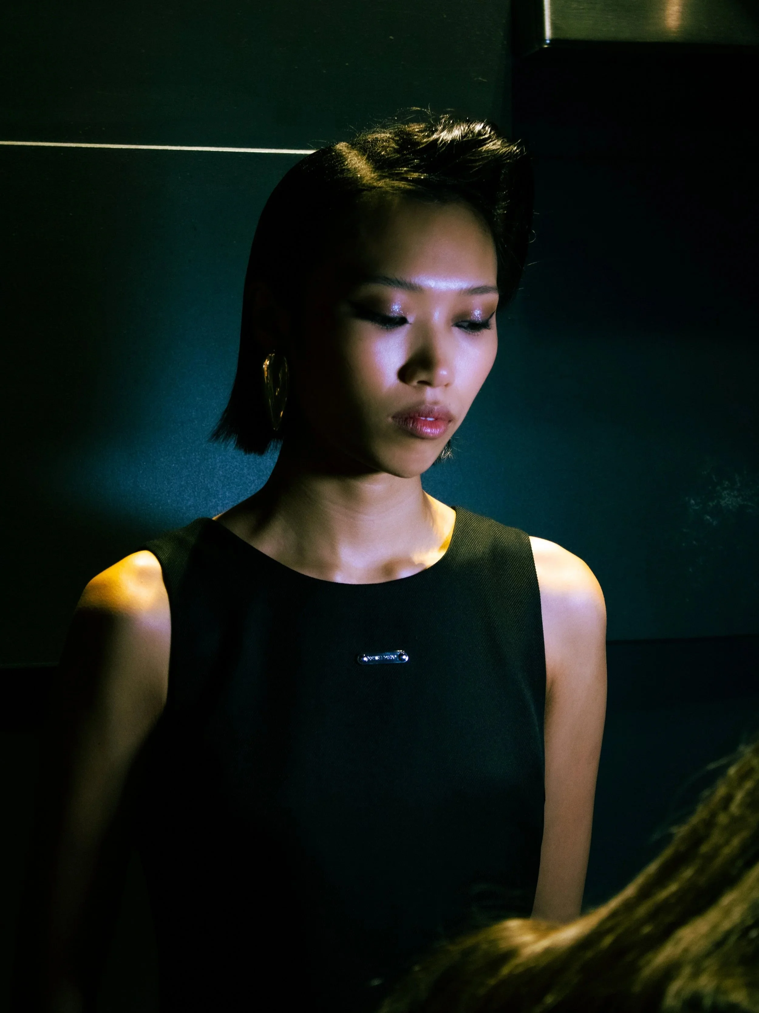 A woman with short black hair wearing a black sleeveless top with gold earrings, looking down in a dimly lit room.