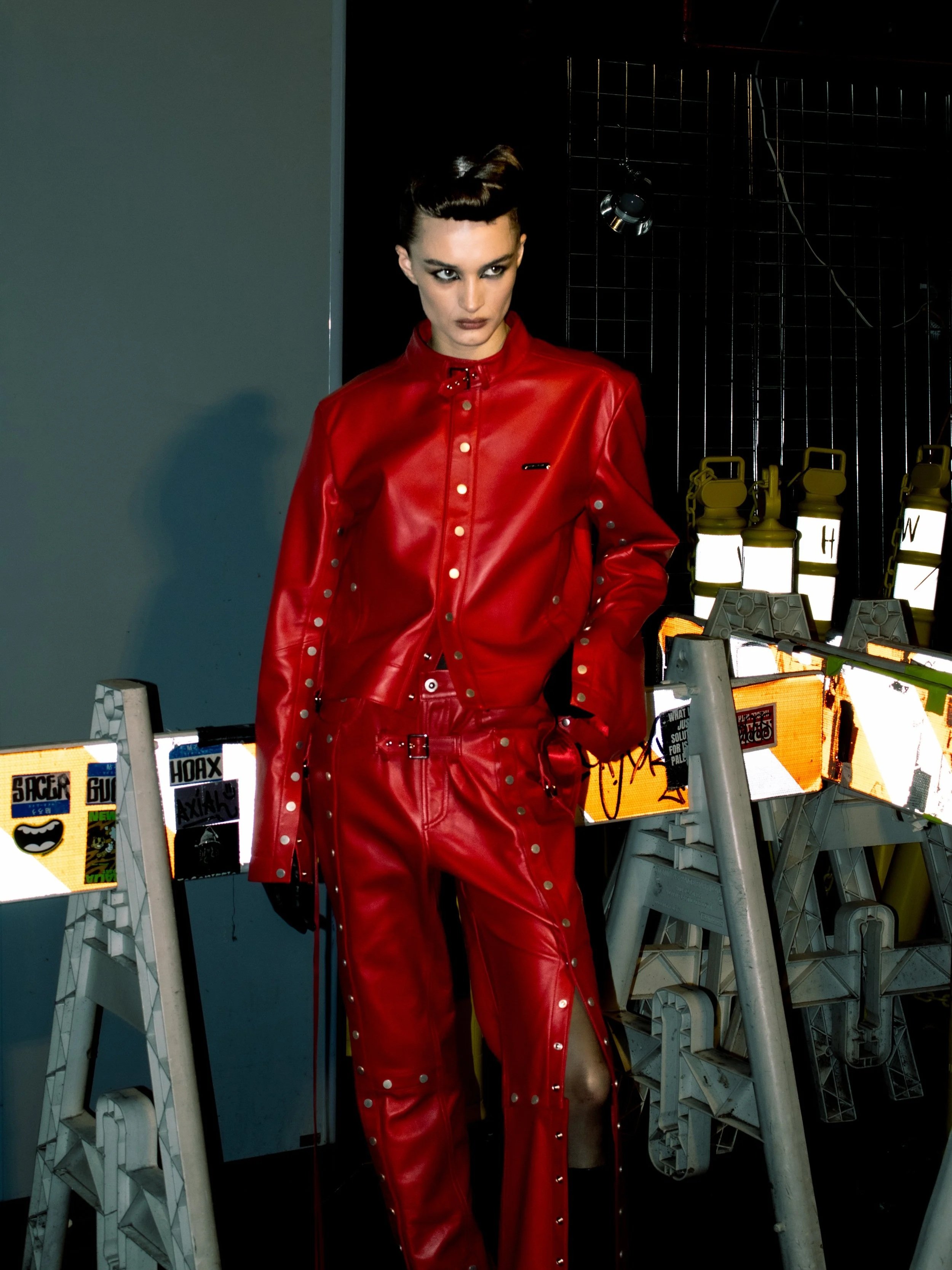 A woman in a shiny red leather jacket and matching red leather pants with metal embellishments, standing in an industrial-like setting with black grid background and illuminated yellow and white signs.