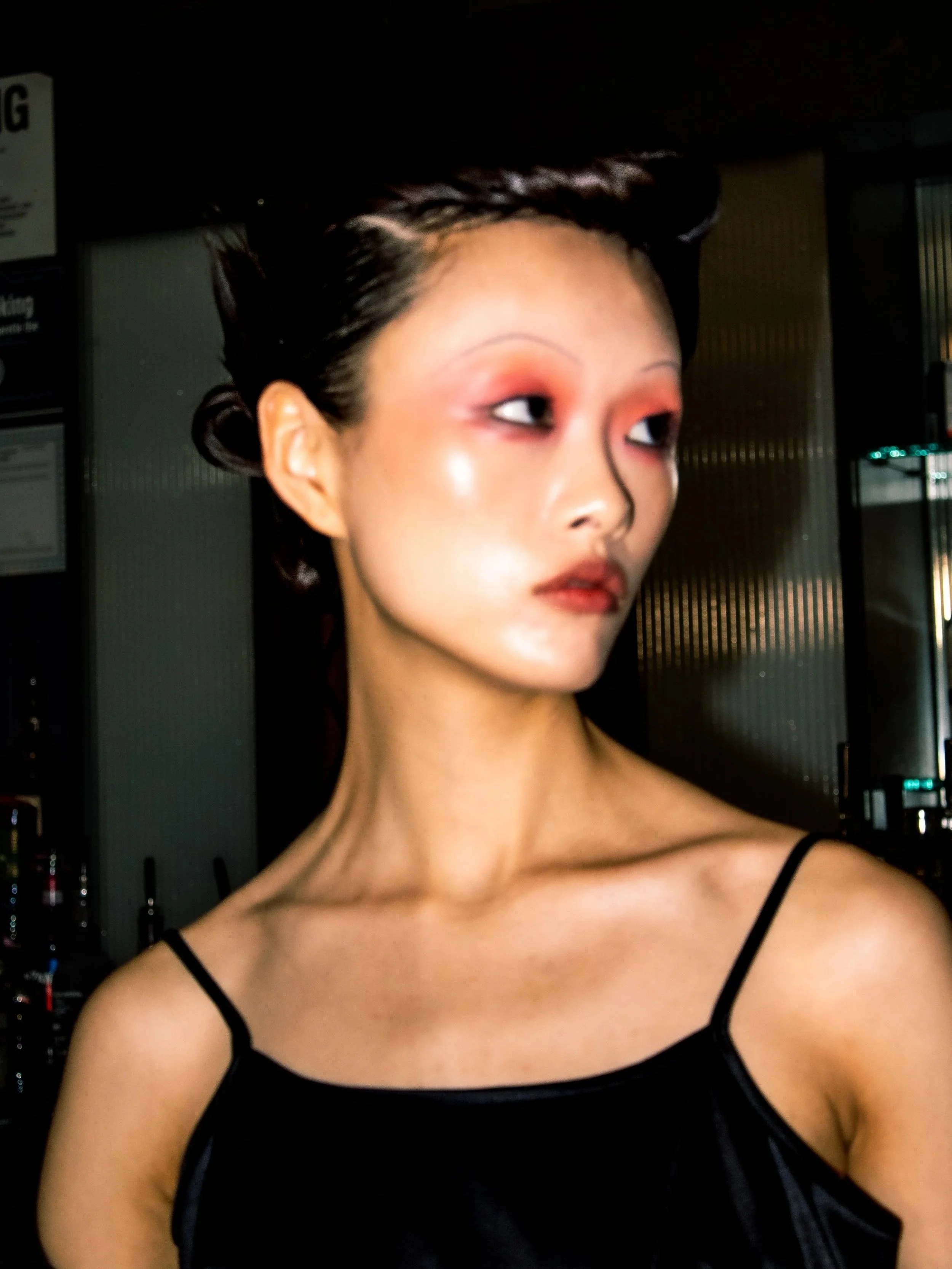 A woman with short dark hair styled in waves, wearing a black spaghetti strap top, looking to her right in an indoor setting with dark background.
