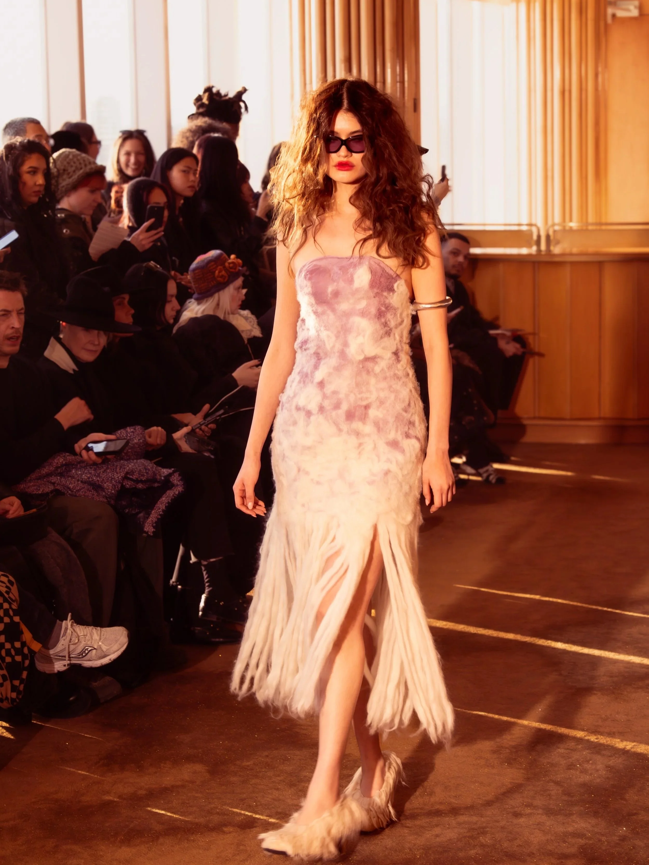 Model walking on runway in a strapless peach and beige dress with fringe details, wearing sunglasses and furry shoes, with an audience seated and watching.