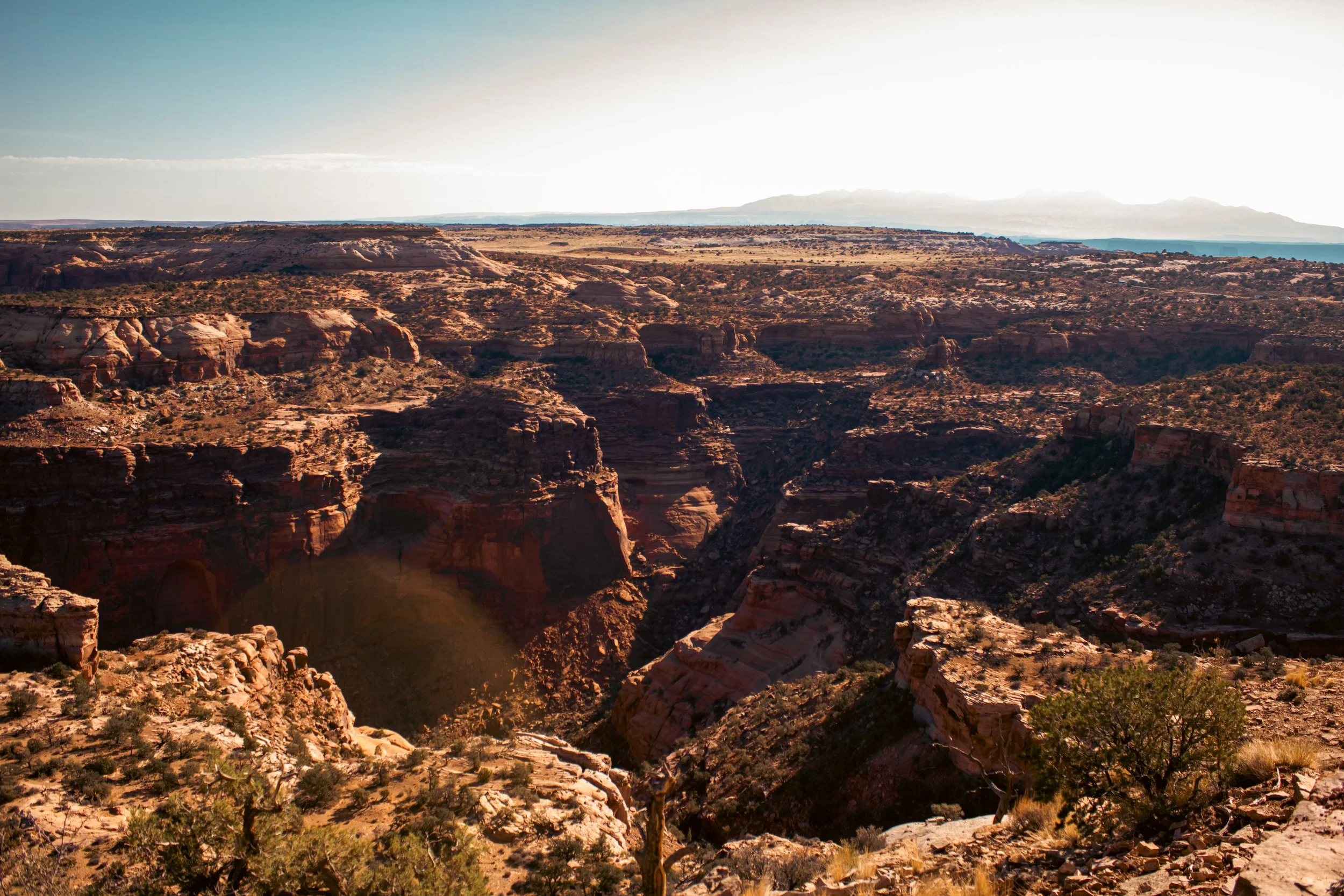 A panoramic view of a rocky canyon landscape with cliffs, sparse vegetation, and a distant horizon under a clear sky.