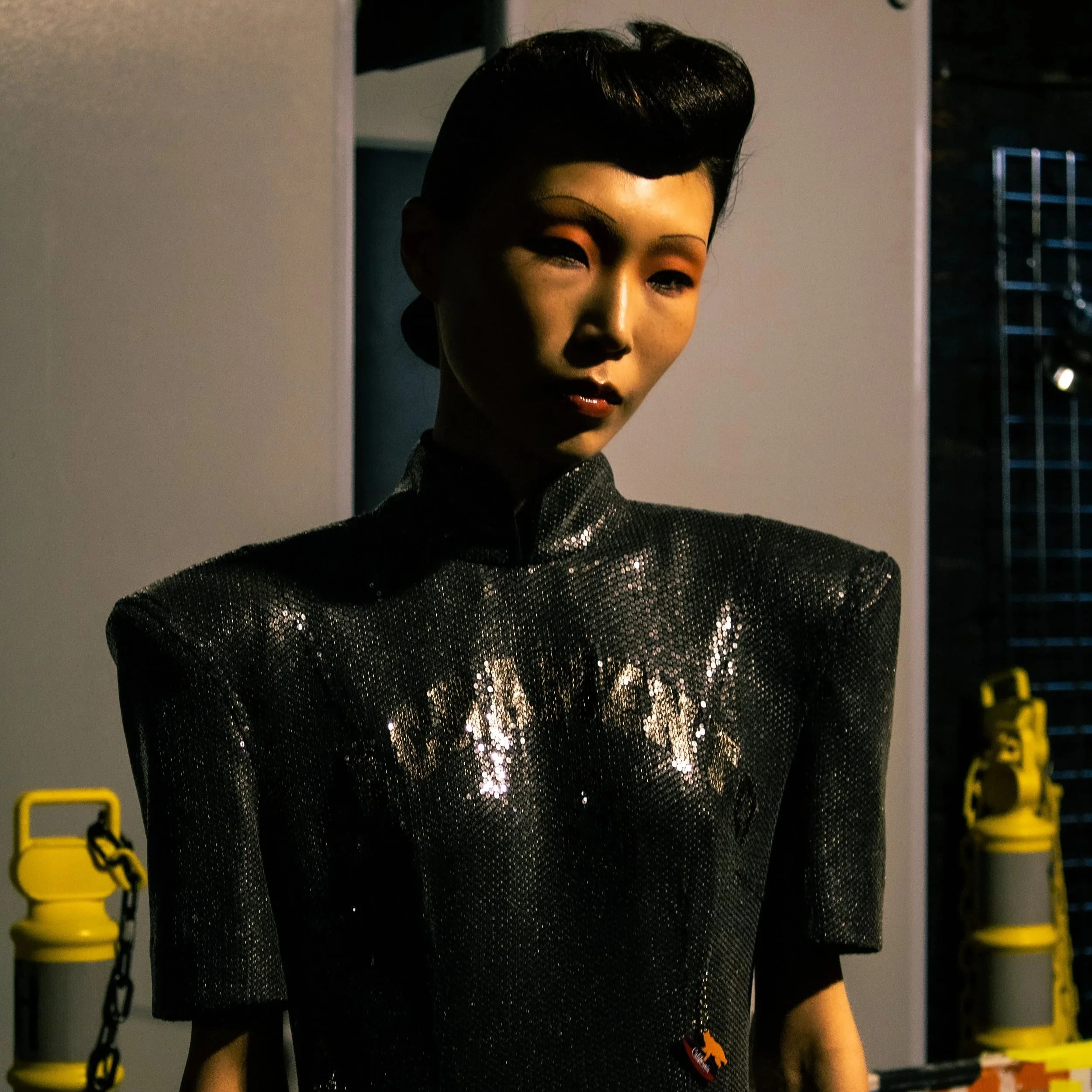 A woman wearing a shiny black, textured top with a high collar in an indoor setting, with yellow chain barriers in the background.