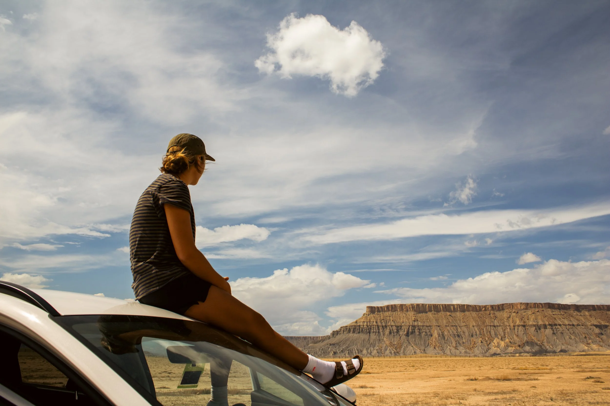 A girl wearing a striped shirt, shorts, sandals, and a cap sitting on the hood of a white car in a desert landscape with mesas and a cloudy sky in the background.