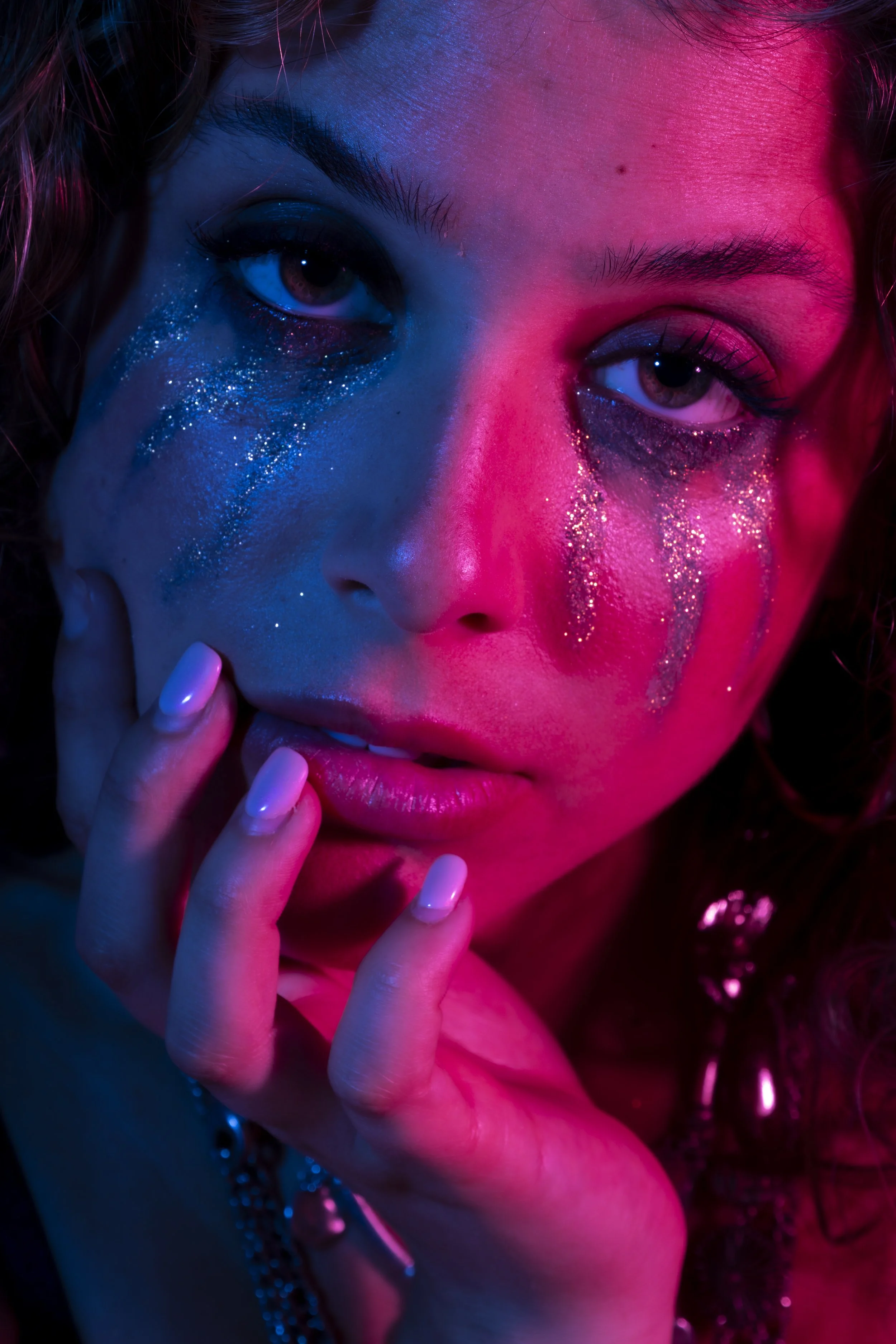 Close-up portrait of a young woman with neon pink and blue lighting, glitter makeup, and hand touching her face.