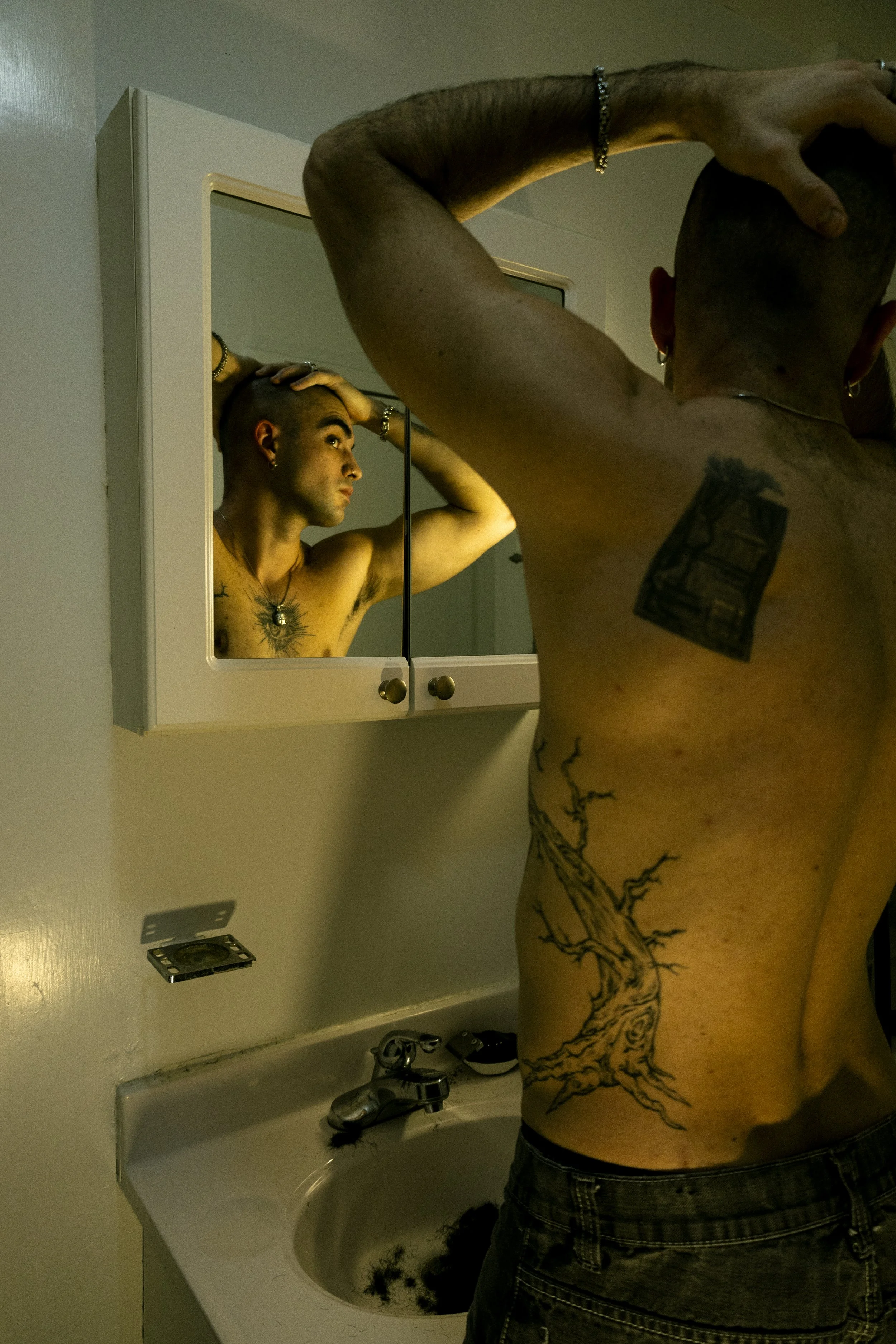 A person with tattoos looking into a bathroom mirror, holding their head with both hands.