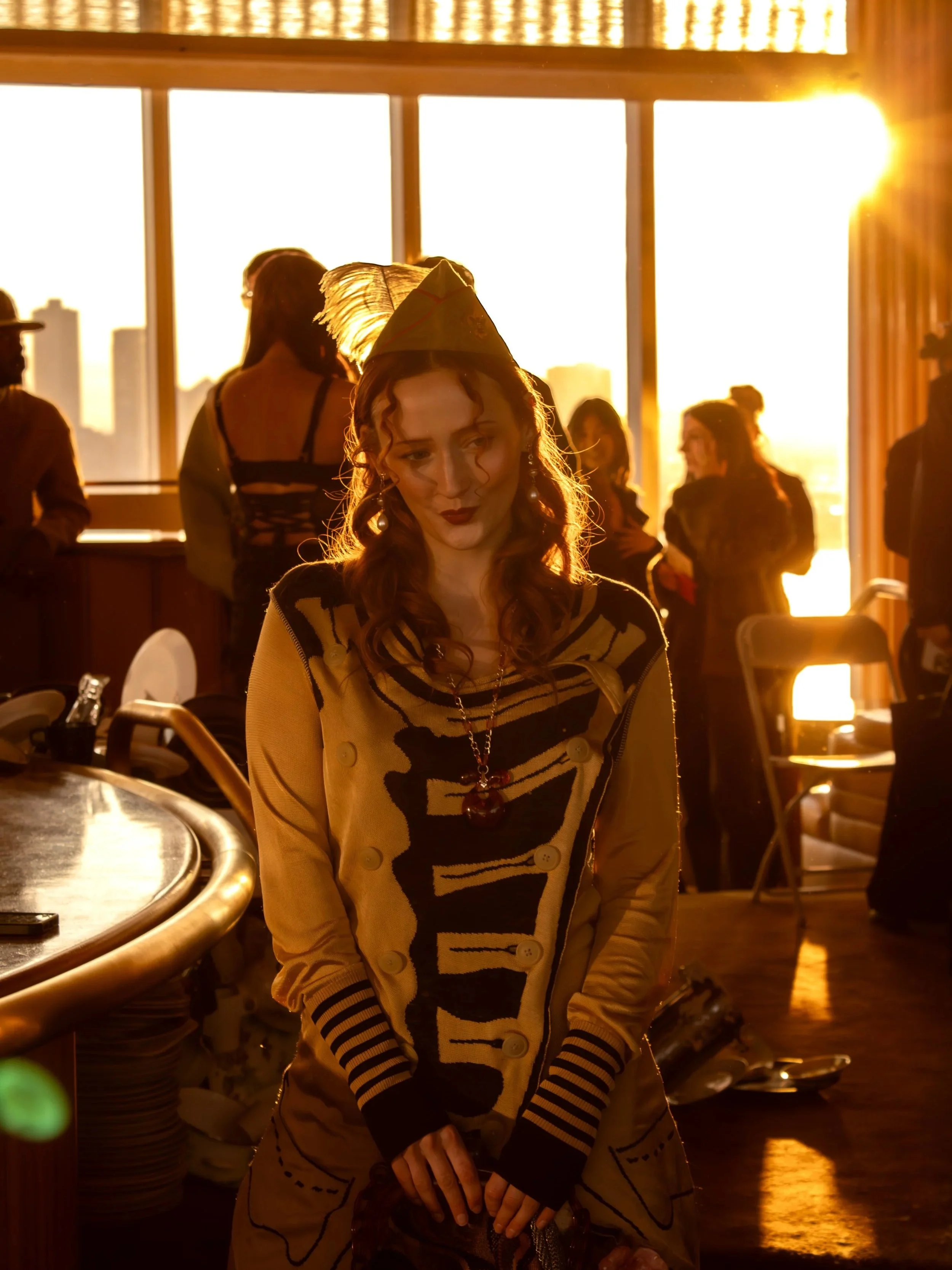 A woman with red hair, dressed in a patterned beige coat and hat, is standing indoors with a sunset view visible through large windows behind her. The room has other people in the background, some standing and some facing the windows, illuminated by the warm glow of the setting sun.