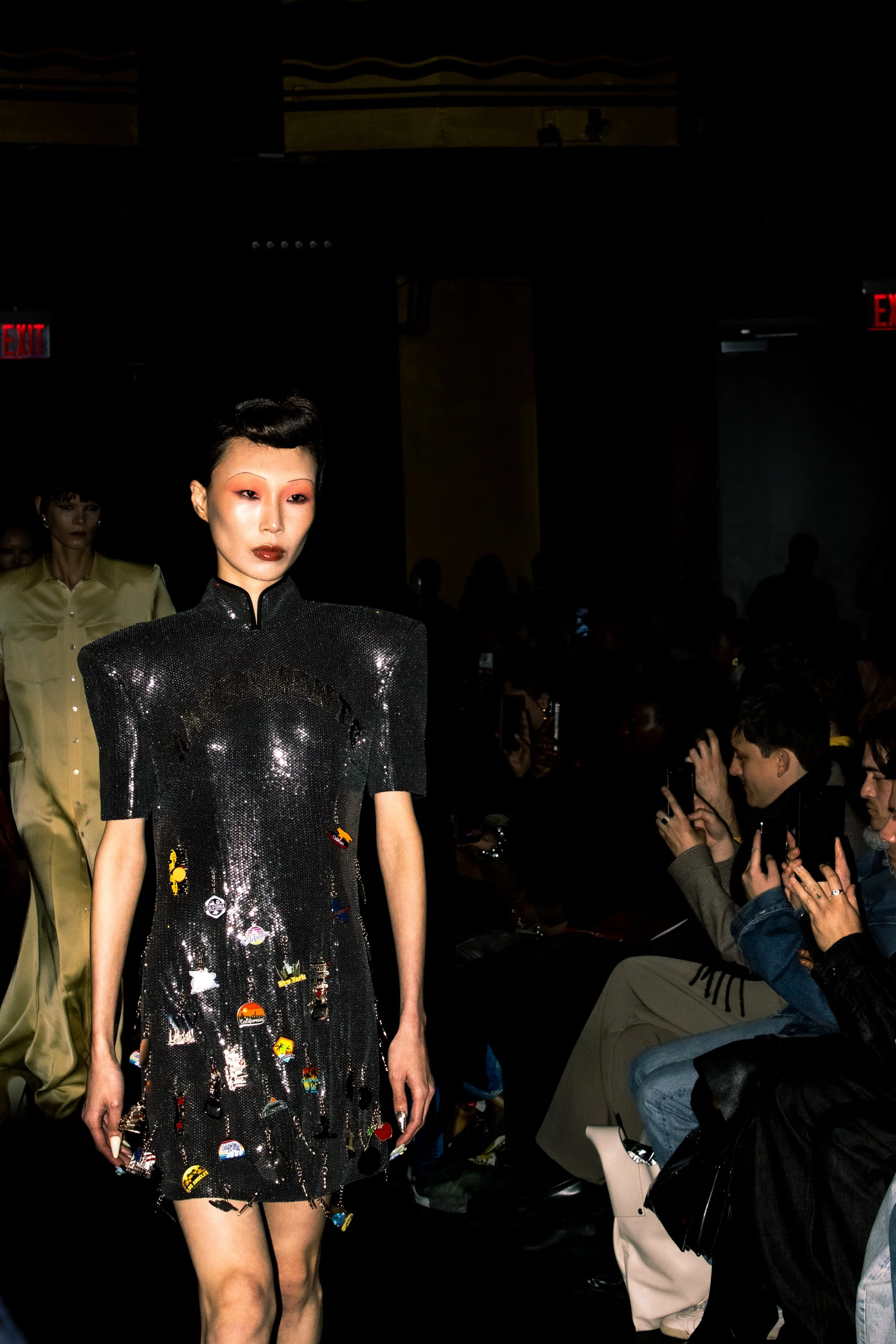 A model walks down the runway in a black, shiny dress adorned with colorful patches at a fashion show. The audience is visible on the right side, with some people taking photographs.