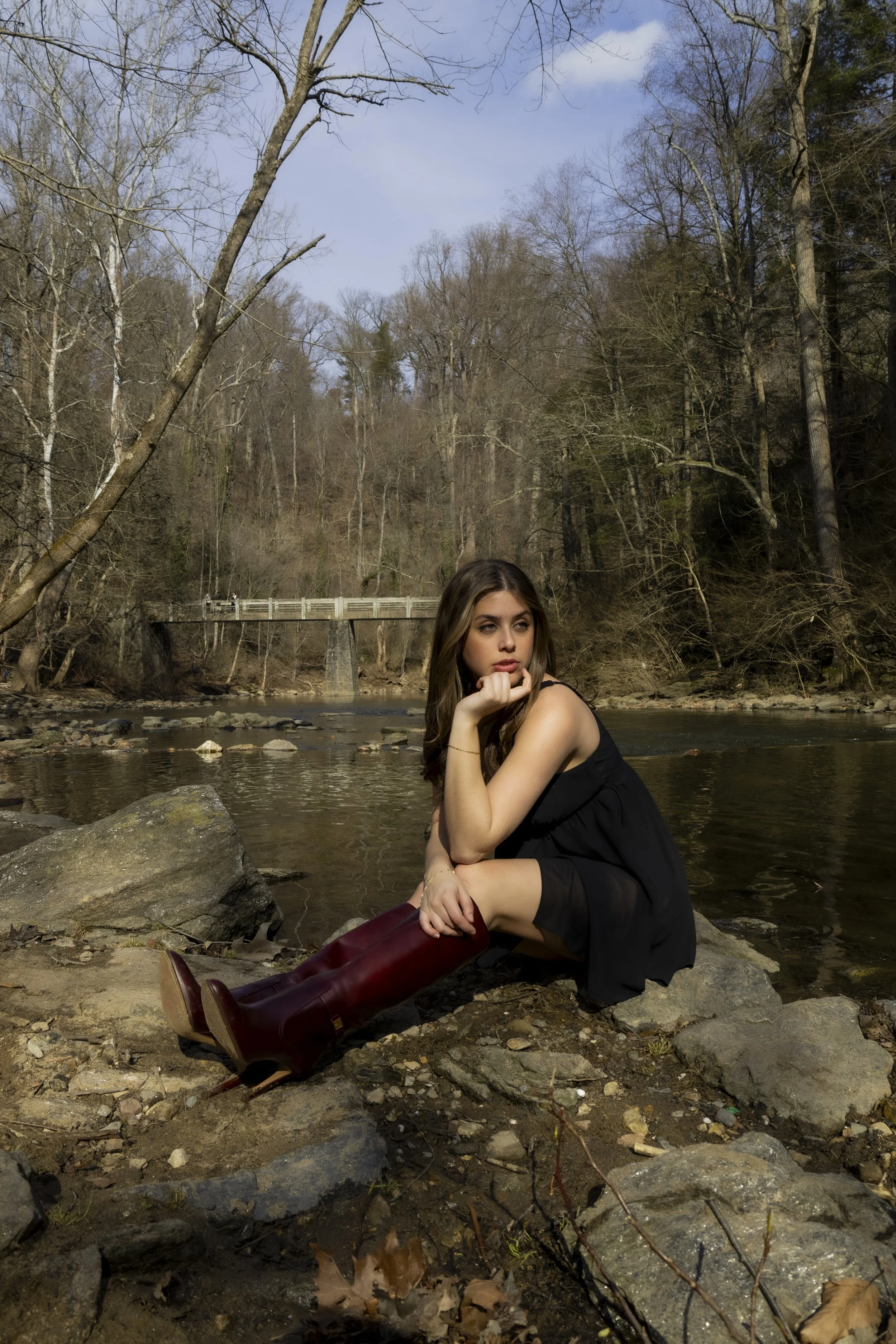 A young woman wearing a black dress and tall maroon boots sitting on rocks near a river in a wooded area with leafless trees and a small bridge in the background.