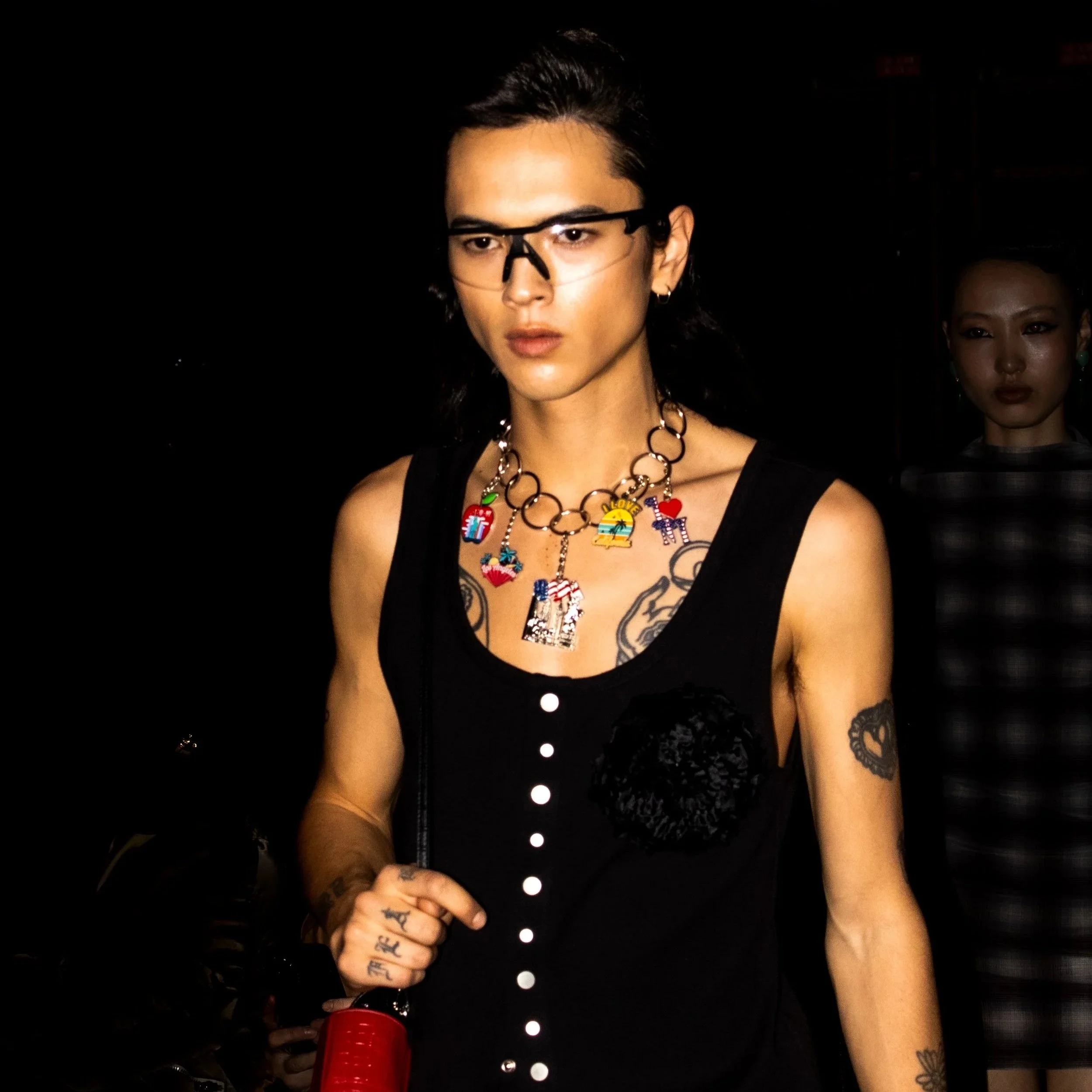 A person with dark hair, wearing black glasses, a black sleeveless top, and colorful charm necklaces, walking in a dark environment down the runway. 