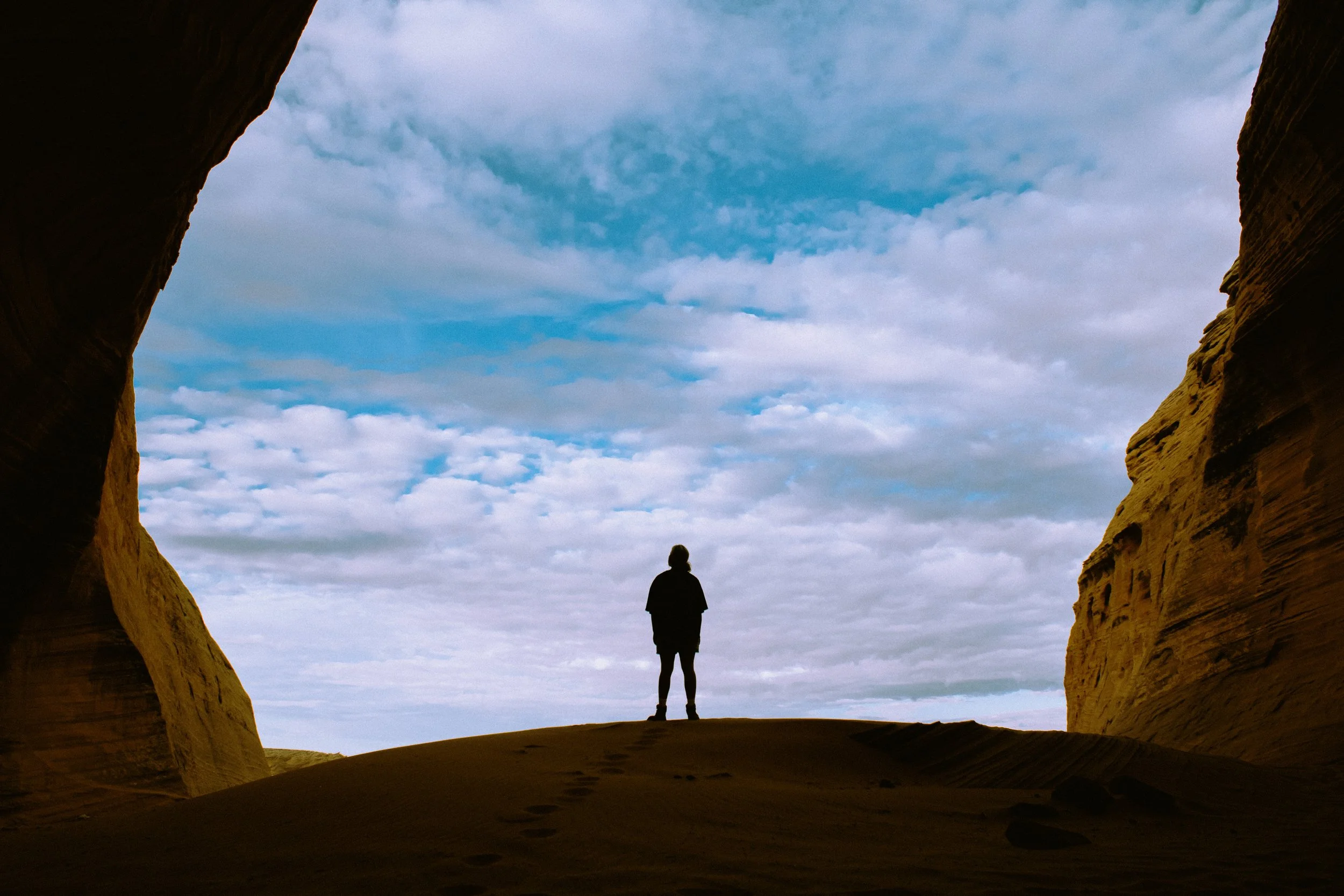 Person standing on sand dune, viewed through a natural rock formation, with cloudy sky in the background.