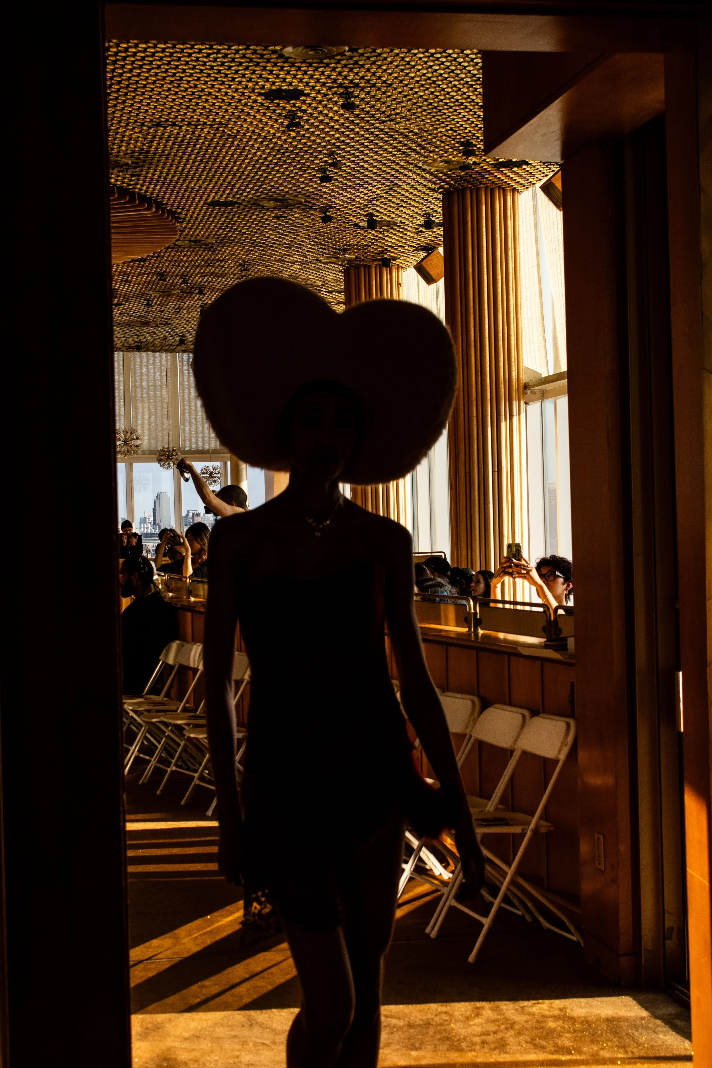 A silhouette of a woman wearing a large heart-shaped headpiece, walking into a room with warm golden lighting, wooden decor, and a patterned ceiling. People in the background are taking photos and sitting near large windows.