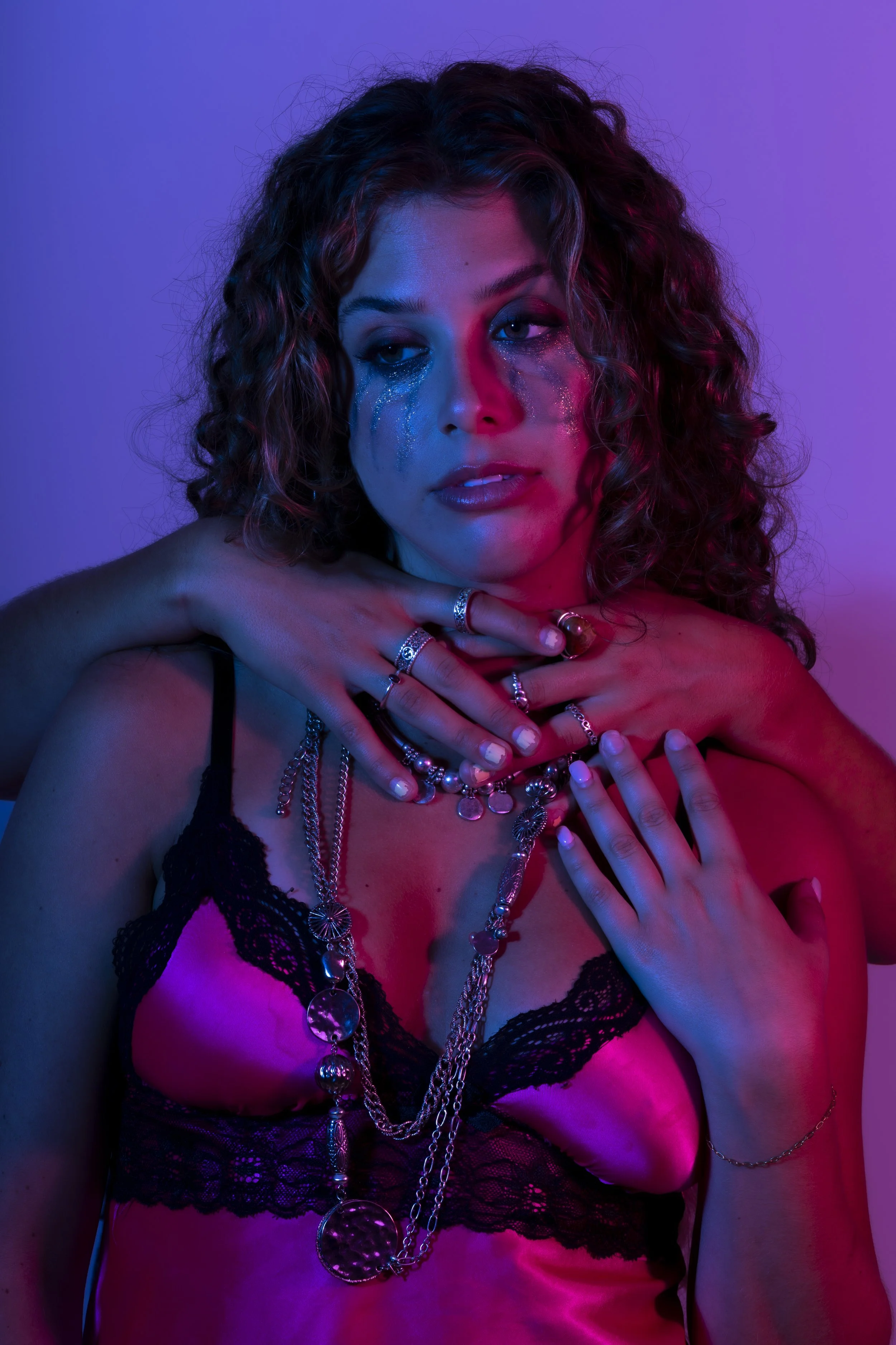 A woman with curly hair is wearing multiple layered necklaces and rings, dressed in a pink satin and black lace top, against a purple background with blue and pink lighting effects.