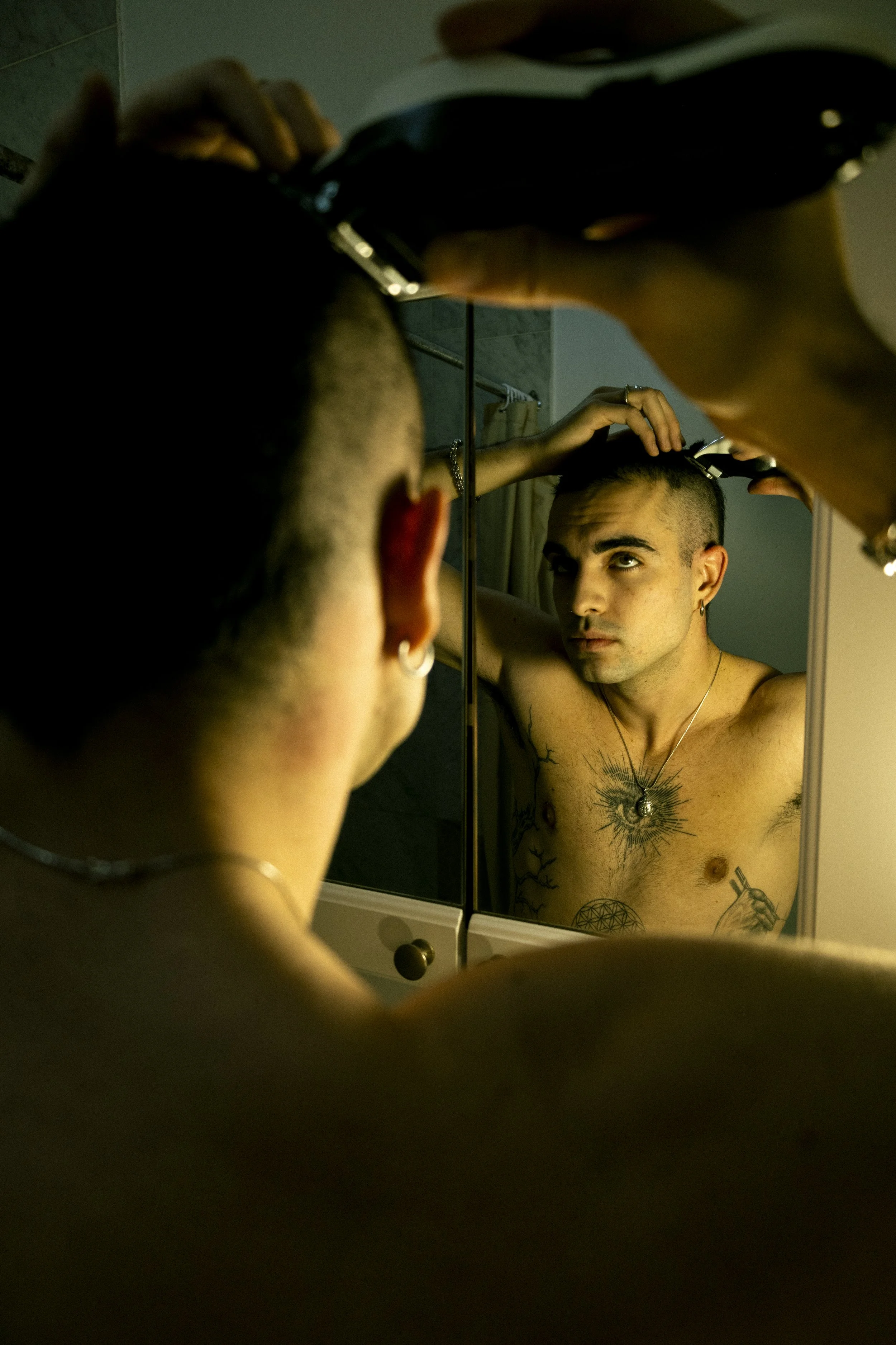 A shirtless man with tattoos, short hair, earrings, and a necklace is looking at himself in the mirror while cutting his hair.
