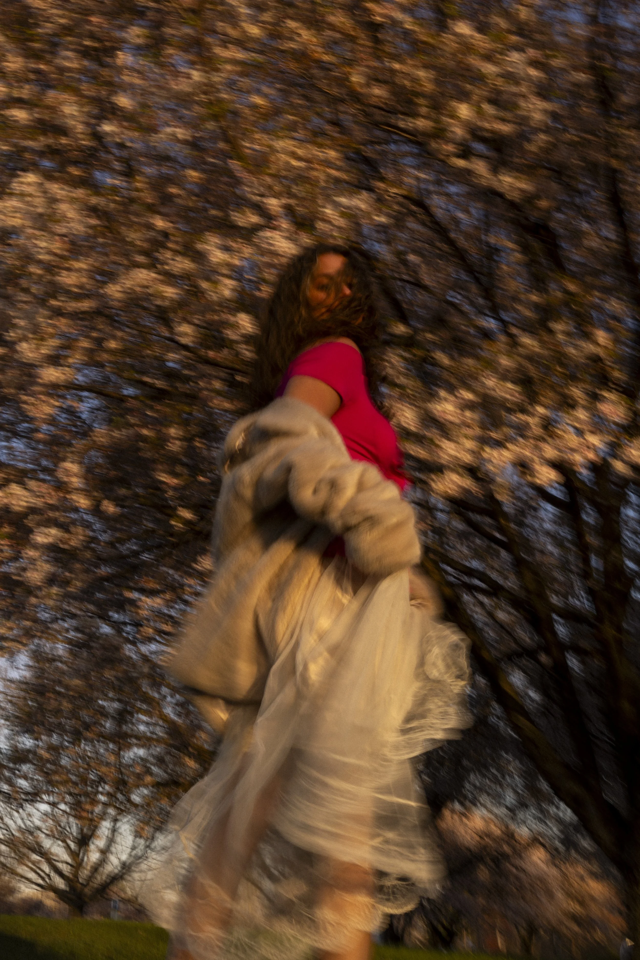 A woman with curly hair wearing a pink top and beige skirt holding onto the edge of her beige coat, standing outdoors in front of a large tree with blooming flowers, during sunset.